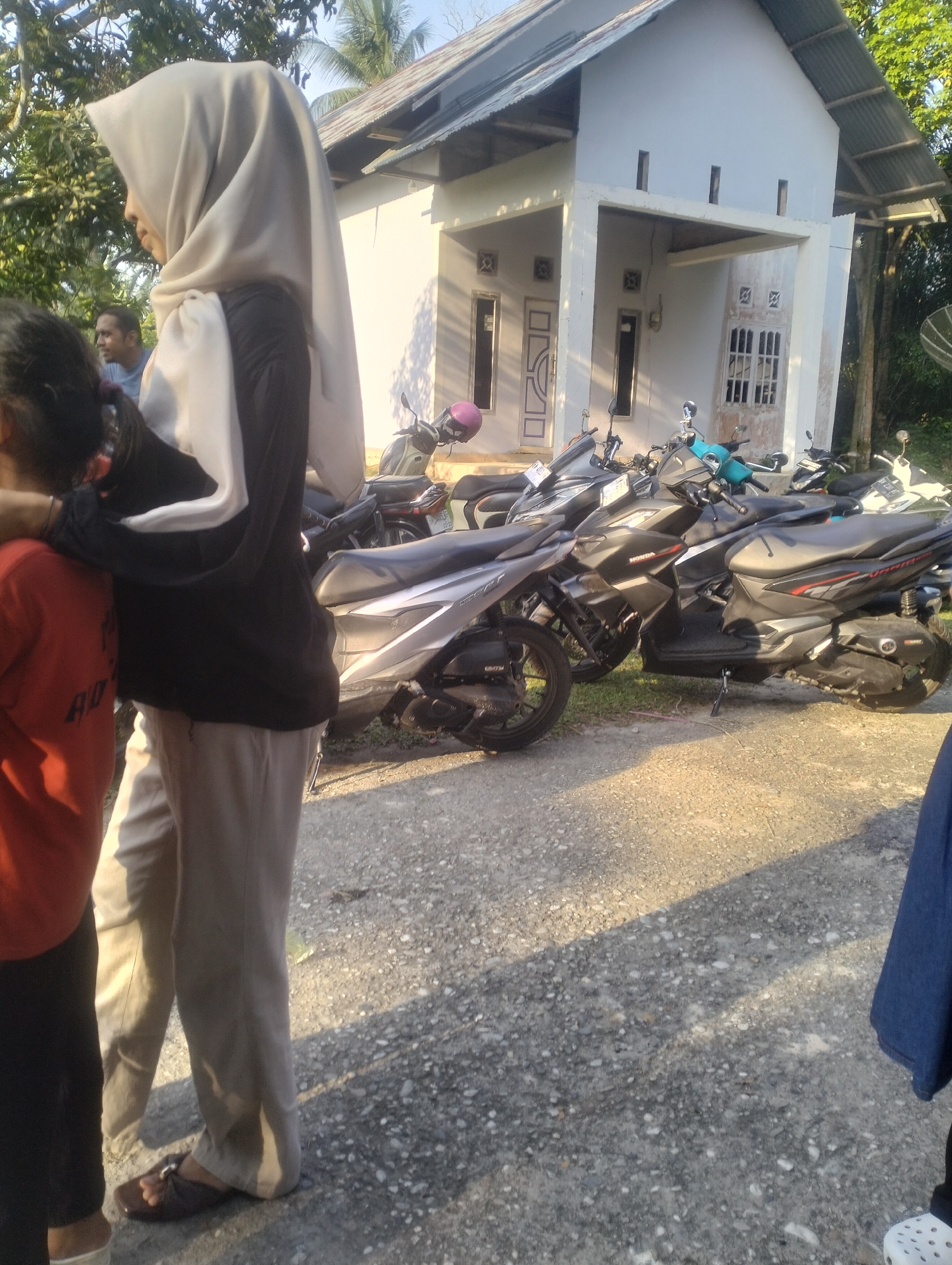 In Dumai City, Indonesia, a woman wearing a light-colored hijab and a black and white long-sleeved shirt stands next to a person wearing a red shirt. The woman is also wearing light-colored pants and dark sandals. Behind them, several motorcycles are parked in front of a white, single-story building with a corrugated metal roof. The building has several windows, and a small covered porch area. The ground is covered in gravel. The scene appears to be outdoors in daylight, with some trees visible in the background.