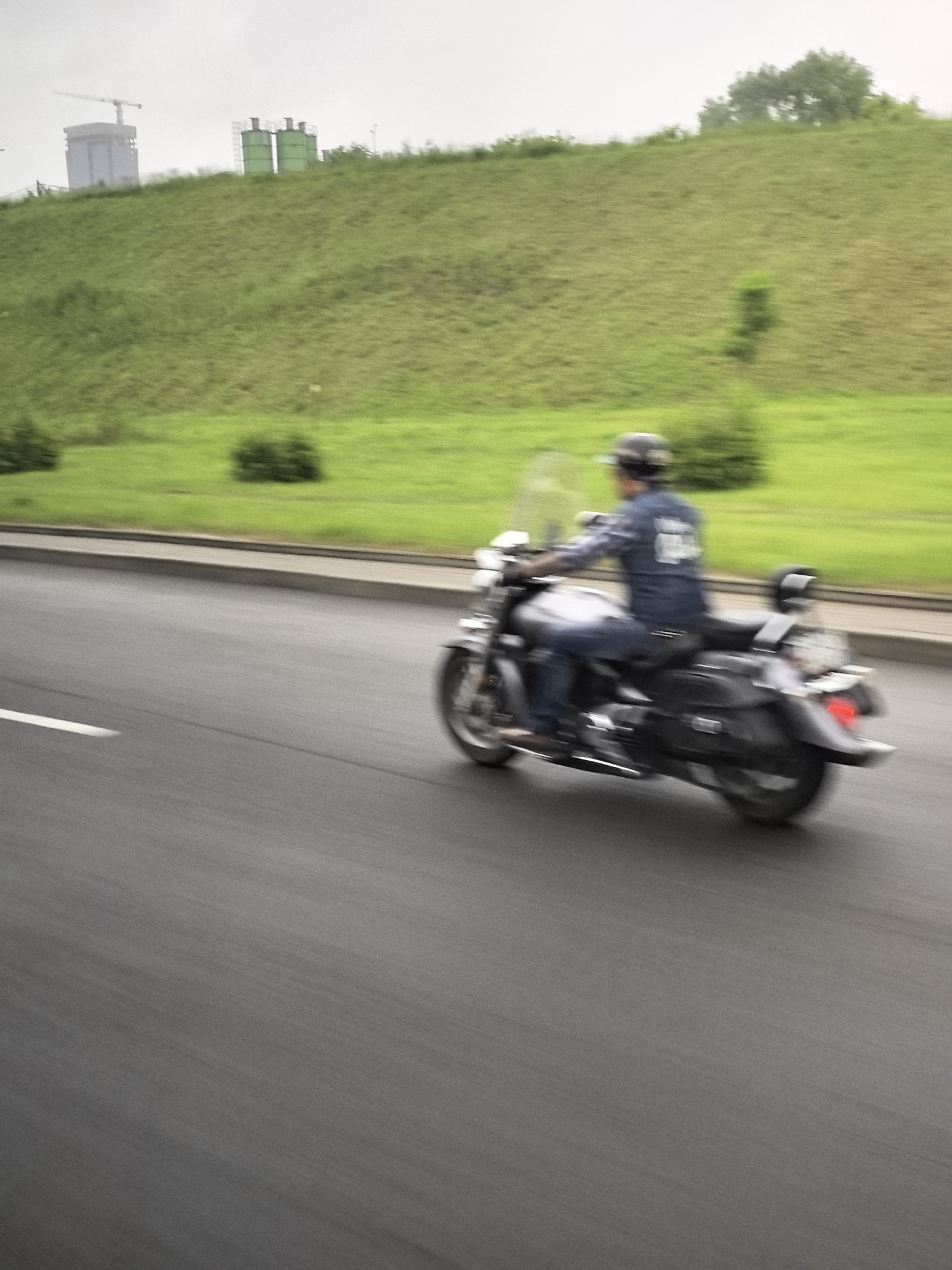 A motion-blurred, eye-level, outdoor shot depicts a person riding a motorcycle on a paved road. The rider, seen from the side and slightly from the back, wears a dark helmet and a dark long-sleeved shirt over a lighter shirt, and dark pants. The motorcycle is dark with a windshield and saddlebags. The road has a white line on the left side. In the background, a green, grassy hillside slopes upward. On the top of the hill, there are some industrial structures, including a tall, light-colored building with a crane and two green cylindrical silos. The sky is overcast and light gray. The image appears to be taken in a suburban or industrial area on an overcast day.
