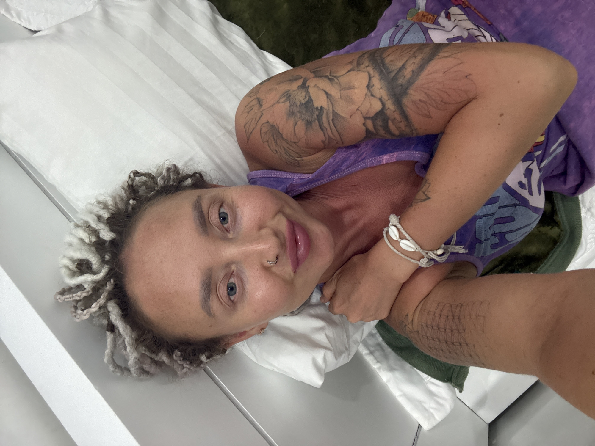 The image shows a woman lying on a bed. She has blonde dreadlocks, blue eyes, and a nose ring. She is wearing a purple tank top and has tattoos on her arms. The bed has white striped sheets and a white pillow. There is a green towel under her head. The background shows a white wall and a dark green carpet. The location is a room in a hotel in the city of Nha Trang, Vietnam.