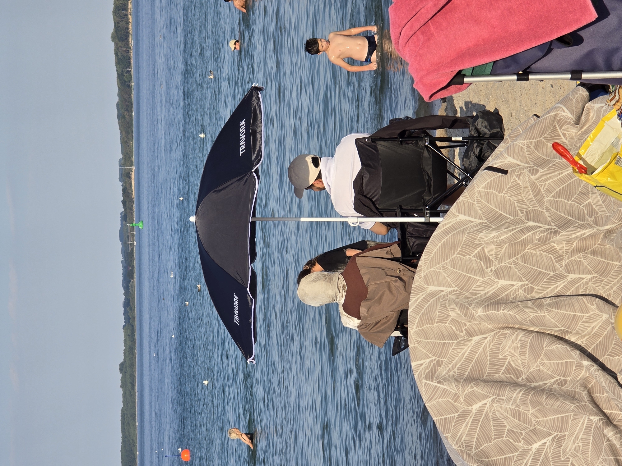 The image depicts a sunny day at the beach with people enjoying the water. In the foreground, a patterned blanket covers a circular object, and a yellow bag with a red ribbon is visible. To the right, a pink towel is draped over a metal frame. Behind these items, two individuals are seated in folding chairs under a black umbrella with the brand name "TRAVORA" visible. One person is wearing a headscarf and a brown and tan outfit, while the other wears a white shirt and a gray baseball cap. In the water, a woman with blonde hair is swimming, and a young boy in black swimming trunks stands in the shallow water. Further in the background, other people are visible in the water, and a tree-lined shore stretches across the horizon. The water is calm, and the sky is clear with a few wispy clouds. The scene suggests a recreational setting, possibly Travemünde Strand, a popular beach area in Lübeck, Germany, known for its sandy shores and recreational activities.