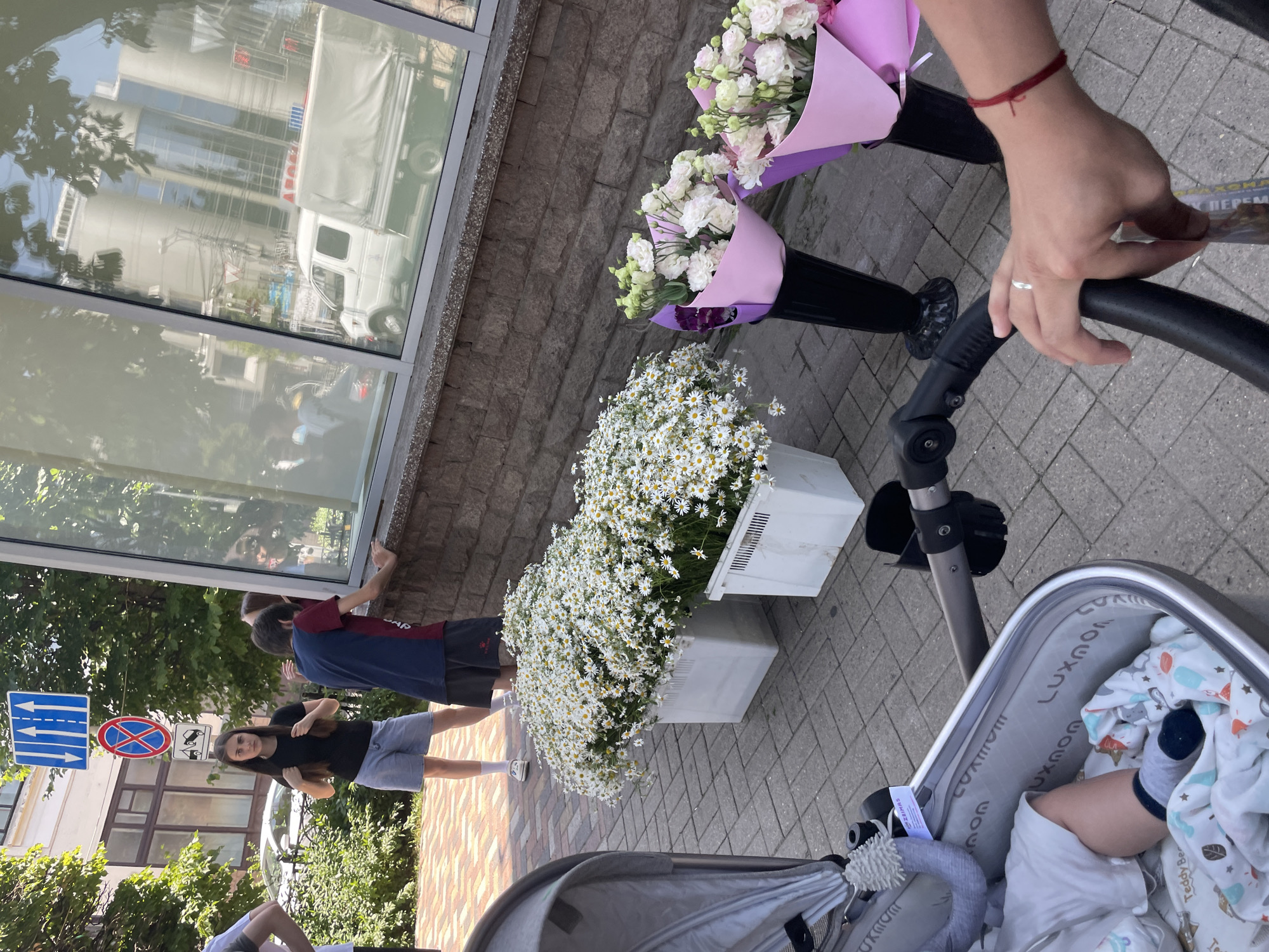 A group of young people are gathered on a paved sidewalk next to a building with large windows. In the foreground, a baby stroller is visible, with a baby's leg and foot sticking out. Two large white crates overflowing with white daisies are placed on the sidewalk. To the right, two bouquets of pink and white flowers are arranged in black vases, wrapped in pink paper. The building's windows reflect trees and a white truck. In the background, a traffic sign with blue arrows and a red circular sign with a diagonal red line are mounted on a pole. One young woman in a black t-shirt and denim shorts stands facing the camera, while another person in a maroon and navy shirt leans towards the window. Another person, partially visible, is also near the window. The environment suggests an outdoor urban setting on a sunny day. The location is identified as Stavropol, Russia.