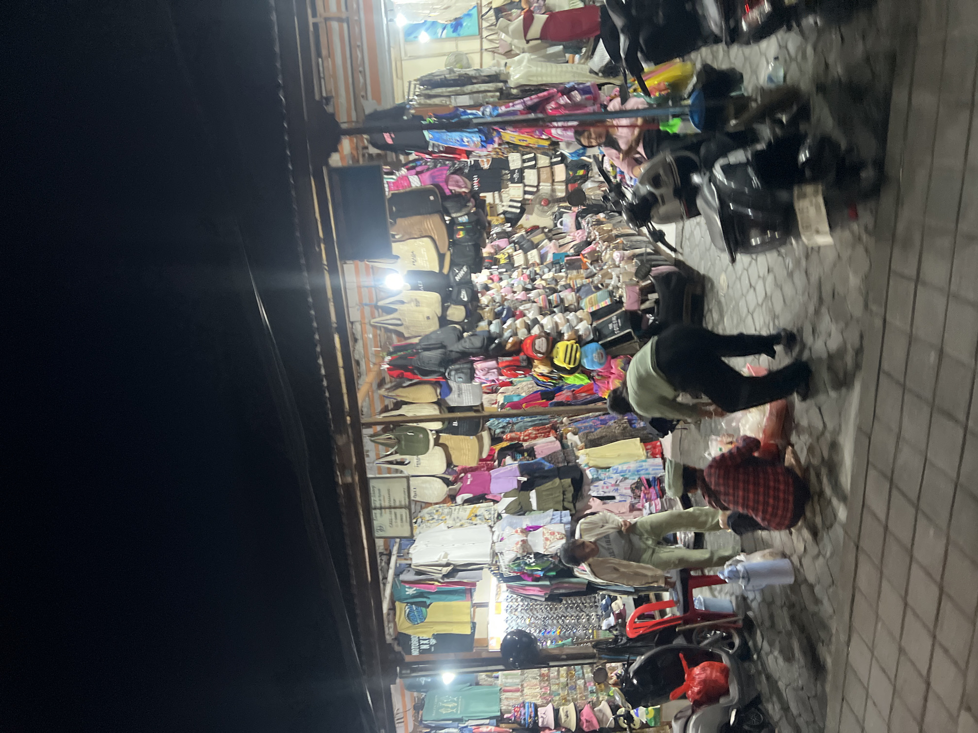 The image depicts a night scene at a market stall in Kuta, Indonesia, characterized by a narrow street or alleyway lined with shops. The stall is densely packed with various items for sale, including clothing, bags, hats, and other accessories. Several people are present, some appearing to be vendors and others possibly customers. Two individuals are crouched down in the foreground, appearing to be interacting with merchandise or packaging. Motorbikes are parked on the side, suggesting transportation. The overall atmosphere is that of a bustling night market.