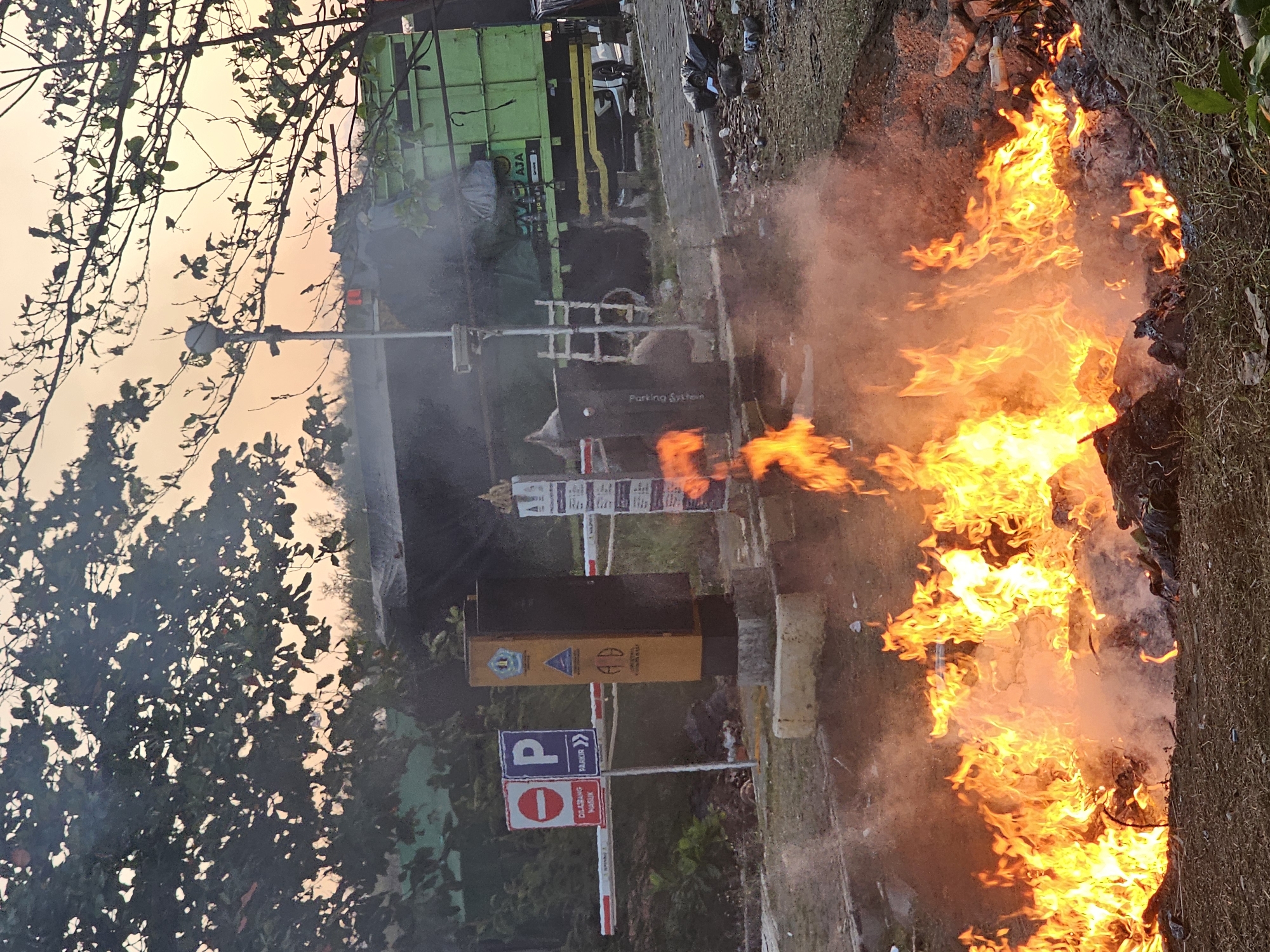 In Denpasar, Indonesia, a controlled burn is underway at what appears to be a refuse collection point or transfer station, indicated by the presence of a large green garbage truck and a barrier system with signage. The foreground is dominated by a significant fire, with flames licking upwards and producing a considerable amount of smoke. The fire is burning what looks to be a pile of organic waste and debris. Behind the fire, a yellow payment or information kiosk stands beside a barrier arm. Signs on the kiosk and a nearby pole include traffic symbols such as a "No Entry" sign and a "P" for parking, suggesting it is an entrance or managed area. A tall light post with a spherical lamp is visible behind the kiosk. In the background, the green garbage truck, possibly a compactor, is positioned, also emitting some smoke. The sky displays an orangish hue, suggesting it might be sunrise or sunset. The general environment appears to be an outdoor, possibly unpaved, area with some vegetation and scattered trash on the ground.