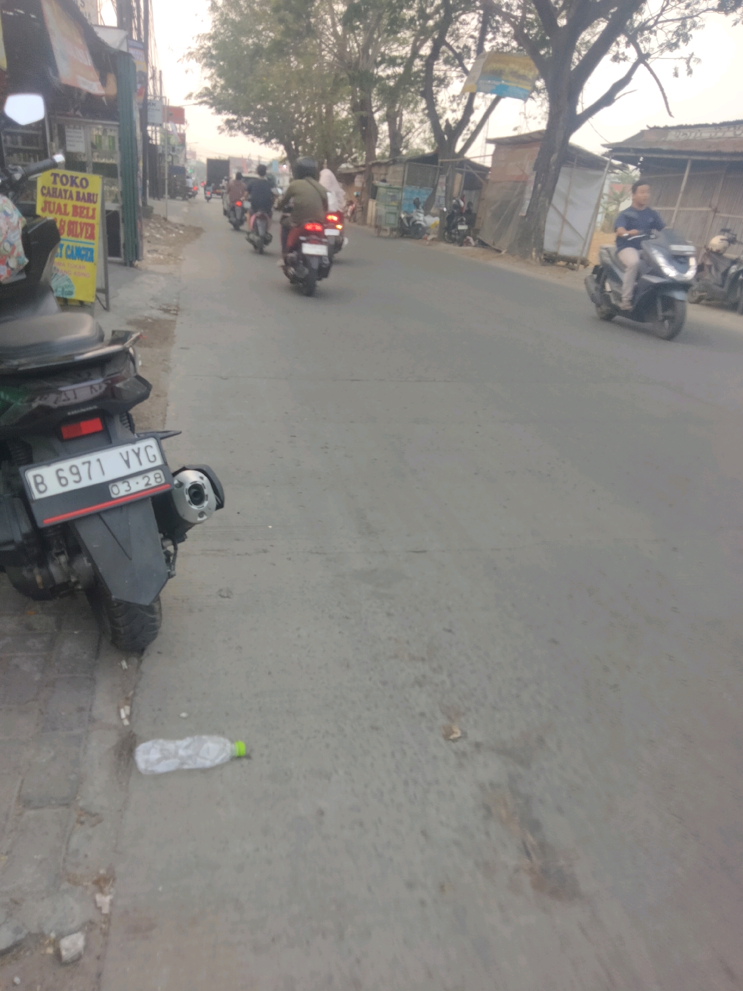 The image depicts a street scene in Sepatan, Indonesia, characterized by a moderate amount of traffic. In the foreground, a motorcycle is parked on the left side of the road, its license plate clearly visible as "B 6971 VYG" with the date "03-28". The road itself is paved and appears to be a typical urban street with shops and buildings lining one side.
Several other motorcycles are in motion on the road, with riders visible on some of them. One motorcycle is traveling towards the viewer and has a rider wearing a dark blue shirt and light-colored pants. Another group of motorcycles is moving away from the viewer, with riders wearing various colored jackets. There are also buildings and trees visible along the sides of the street, contributing to the urban environment. A plastic water bottle is lying on the ground near the parked motorcycle. The overall lighting suggests it is daytime, possibly late afternoon, given the warm tones in the sky. The scene conveys a sense of everyday activity and transportation within the Indonesian city.