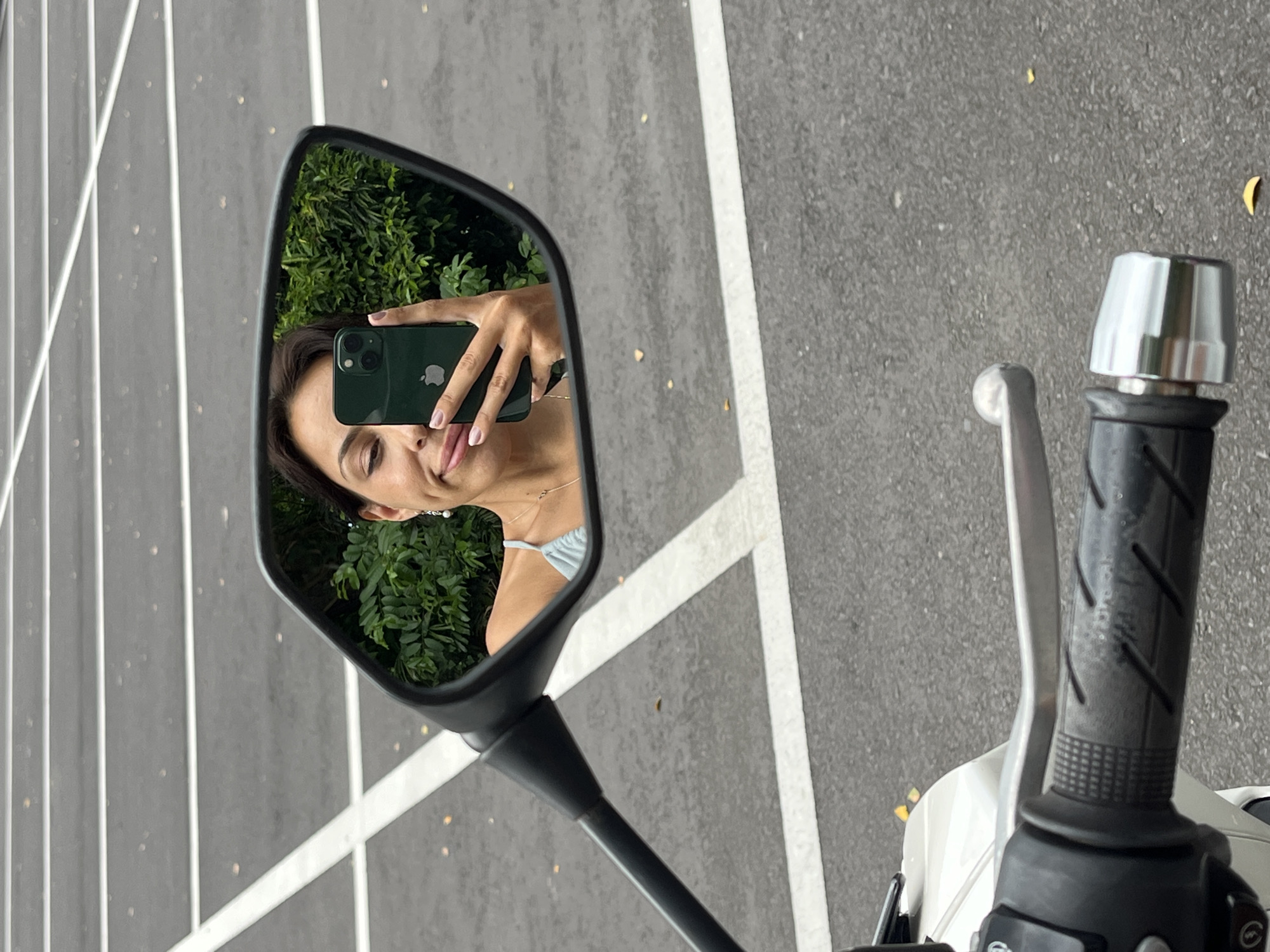 An outdoor, eye-level shot captures a selfie being taken in the side mirror of a motorcycle. The central focus is the motorcycle's black-framed mirror, reflecting a person, likely female, taking the photo. Her face, partially visible, shows a faint smile. She holds a dark green iPhone with a visible Apple logo and dual cameras in her right hand, positioned to capture her reflection. She is wearing a light blue top with thin straps and has a delicate necklace and an earring.
The background reflected in the mirror consists of vibrant green foliage, suggesting lush vegetation. Outside the mirror's reflection, the scene is dominated by dark asphalt pavement with distinct white painted lines, indicative of a parking area or road markings. Part of the motorcycle's handlebar and grip is visible on the right side of the image, featuring a black textured rubber grip, a silver brake/clutch lever, and a silver end cap. The motorcycle itself appears to be white or light-colored in the visible section. The overall lighting suggests it is daytime. The combination of green foliage and pavement aligns with the provided location context of Kuta Selatan, Indonesia.