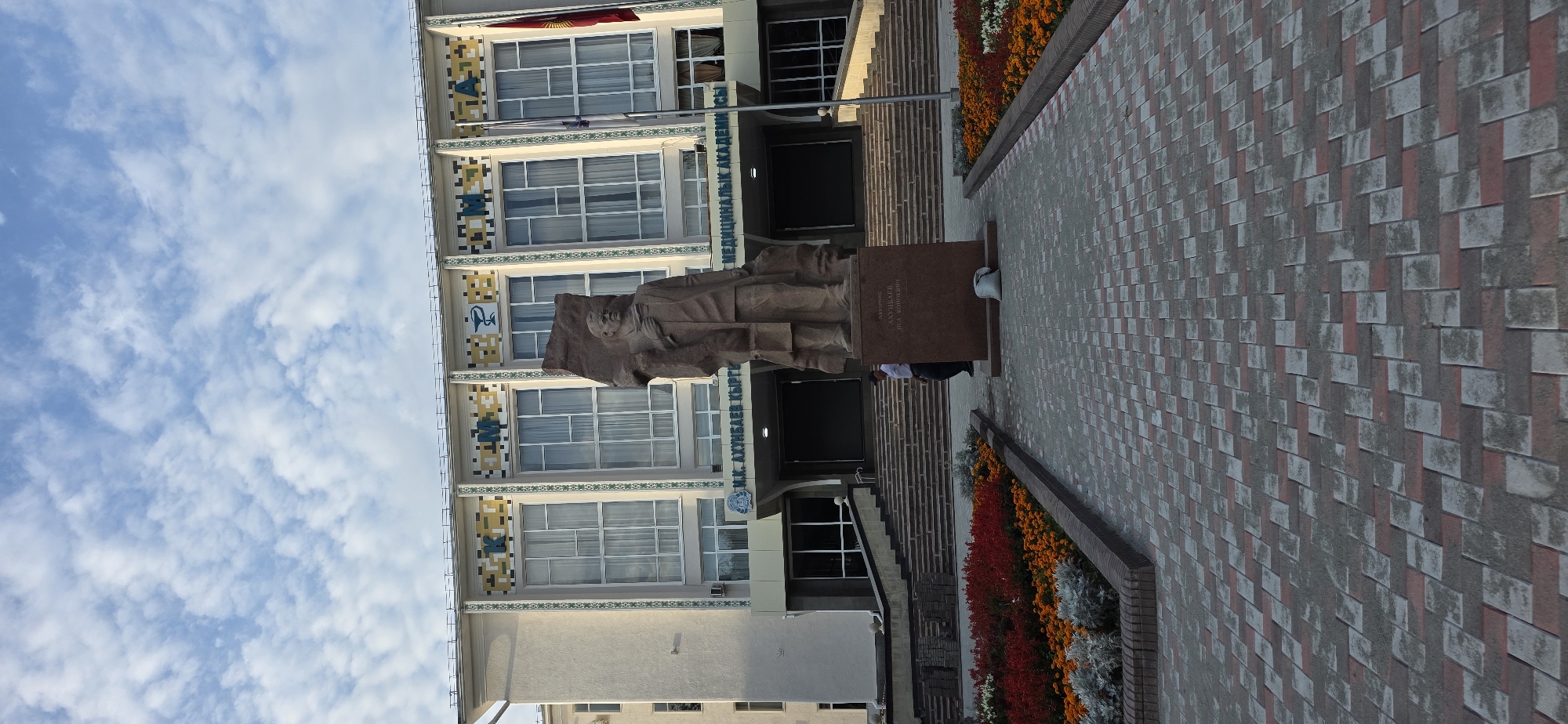 In front of the Kyrgyz State Medical Academy building in Bishkek, Kyrgyzstan, a large stone statue stands on a pedestal. The statue depicts a man in formal attire holding a book. The building behind the statue has a light-colored facade adorned with decorative tiles and large windows. A paved walkway with red and gray bricks leads towards the entrance, with flower beds filled with red and orange flowers on either side. A person in a white shirt and dark pants is visible walking on the stairs to the left of the statue, and a sandbag is placed at the base of the pedestal. The sky above is partly cloudy. The inscription on the pedestal appears to be in Cyrillic.