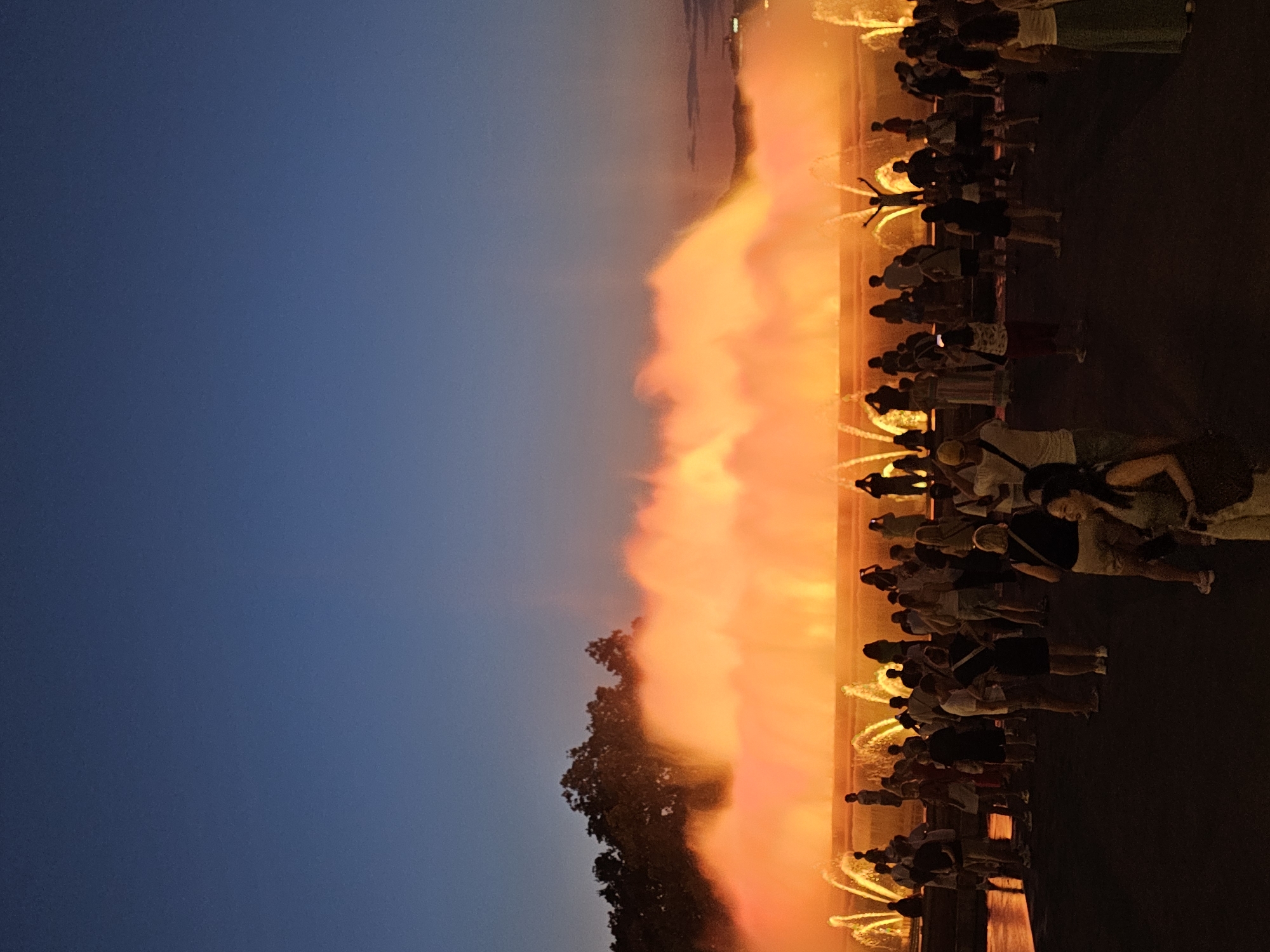 The image depicts a view of the Magic Fountain of Montjuïc in Barcelona, Spain, during the evening. A large crowd of people is gathered in front of the illuminated fountain, which is spraying water in vibrant orange and yellow hues against a darkening blue sky. The fountain's lights create a dramatic and colorful display, with water plumes rising high into the air. Silhouettes of trees are visible in the background, silhouetted against the bright fountain. The foreground shows a variety of individuals and small groups of people standing and observing the spectacle. Some are holding up phones, likely to capture photographs or videos of the fountain's performance. The ground in the foreground appears to be a dark, paved surface.
