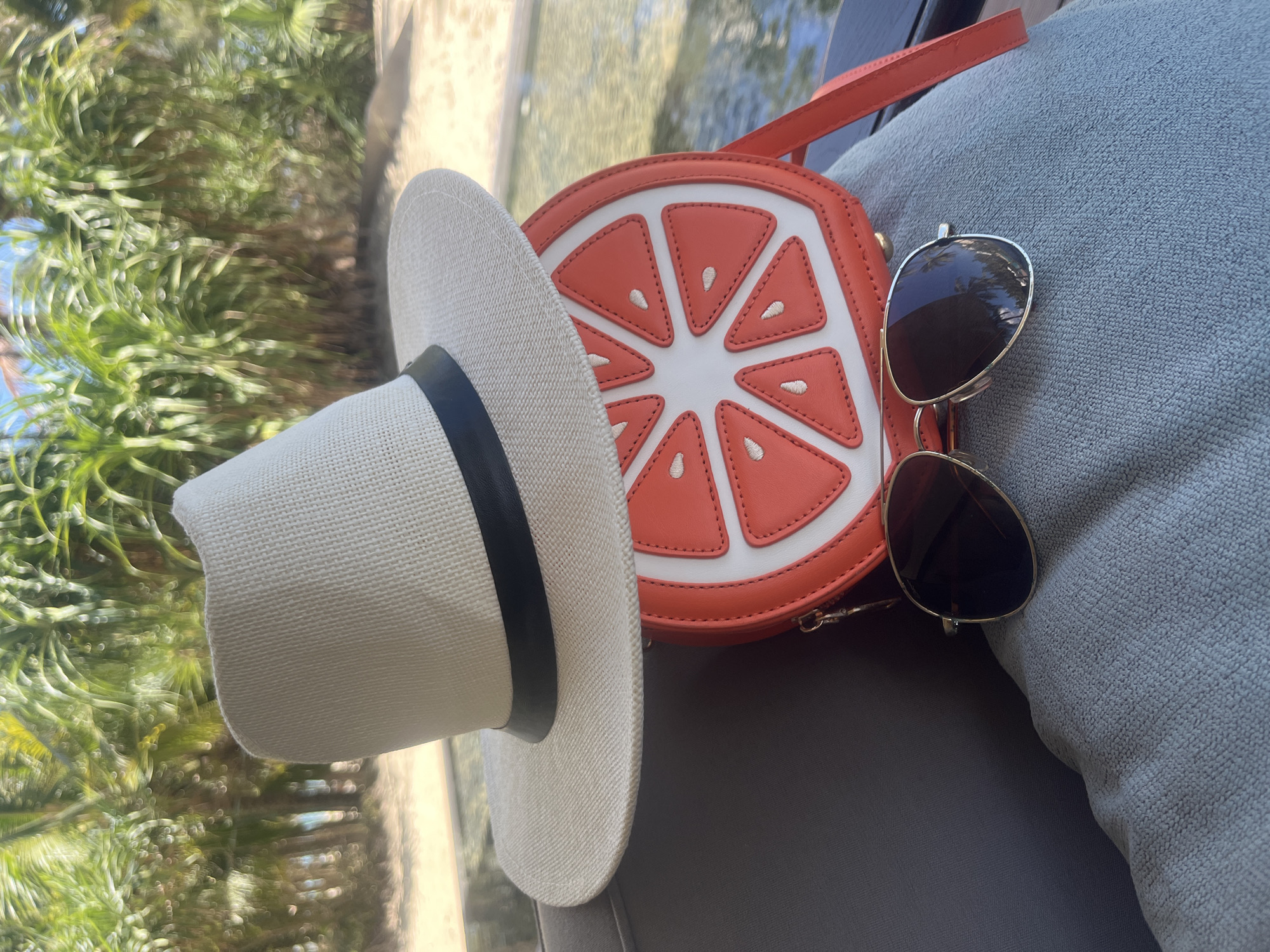This image displays a collection of summer accessories resting on a grey, textured surface, likely an outdoor cushion or lounge chair, in what appears to be a tropical setting like Đà Nẵng, Vietnam. The primary subjects include a light-colored straw panama hat with a black band, an distinctive orange and white circular handbag designed to resemble a grapefruit slice, and a pair of gold-framed aviator sunglasses with dark lenses.
In the background, lush green foliage, consistent with palm trees or other tropical plants, is softly blurred, indicating an outdoor environment under bright sunlight. Hints of shimmering water and a light-colored surface, possibly sand or a path, are also visible, suggesting a beachside, poolside, or resort location.
The scene takes place during bright daylight, indicated by the clear illumination and lack of harsh shadows. The items evoke a sense of leisure, vacation, and relaxation, suitable for warm, sunny weather. No specific activity is being performed, but the arrangement of the objects suggests readiness for a day out or unwinding after outdoor activities. There is no visible text present in the image.