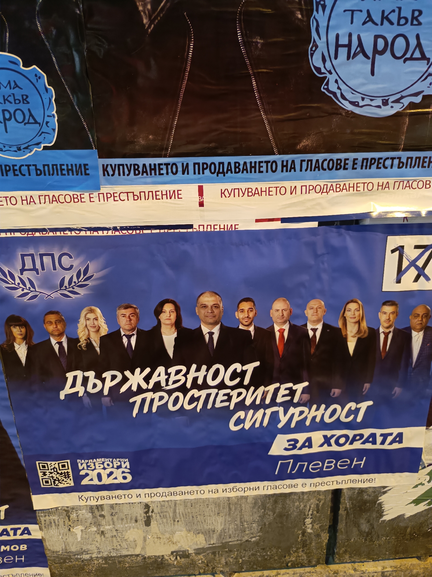 The image shows a Bulgarian political poster on a wall. The poster promotes the DPS party for the parliamentary elections in 2026. It features a group photo of party members and the slogan "STATEHOOD, PROSPERITY, SECURITY FOR THE PEOPLE of Pleven." The number "17" is also prominently displayed. Other posters are visible on the wall above, including one with the phrase "There is such a nation." Several of the signs state, "Buying and selling votes is a crime!" The background and surrounding wall appear weathered and urban.