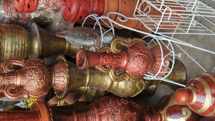 This image captures a dense display of pottery and decorative items at an outdoor vendor stall in Dhaka, Bangladesh. The scene is dominated by a variety of ornate ceramic and terracotta objects, predominantly in shades of metallic copper-bronze, gold, and natural reddish-brown.
In the center, a prominent amphora-shaped vase, painted in a shimmering copper-bronze with gold accents on its two handles and raised floral motifs, rests on a white metal tiered stand. To its right, a silver-painted fish sculpture, detailed with scales and fins, occupies another white metal stand. Directly behind these, several tall, intricately patterned vases are stacked, some featuring horizontal bands and relief floral designs in gold and bronze.
On the far right, stacks of smaller, reddish-brown terracotta lidded containers with geometric cut-out patterns are visible. In the lower left foreground, a reddish-brown bell-shaped piggy bank with golden stripes and a coin slot is partially visible. Additional white metal display racks, some multi-tiered, are interspersed throughout the display. The ground surface appears to be a light-colored, possibly concrete or tiled, outdoor pavement. No people or active interactions are present in the frame.