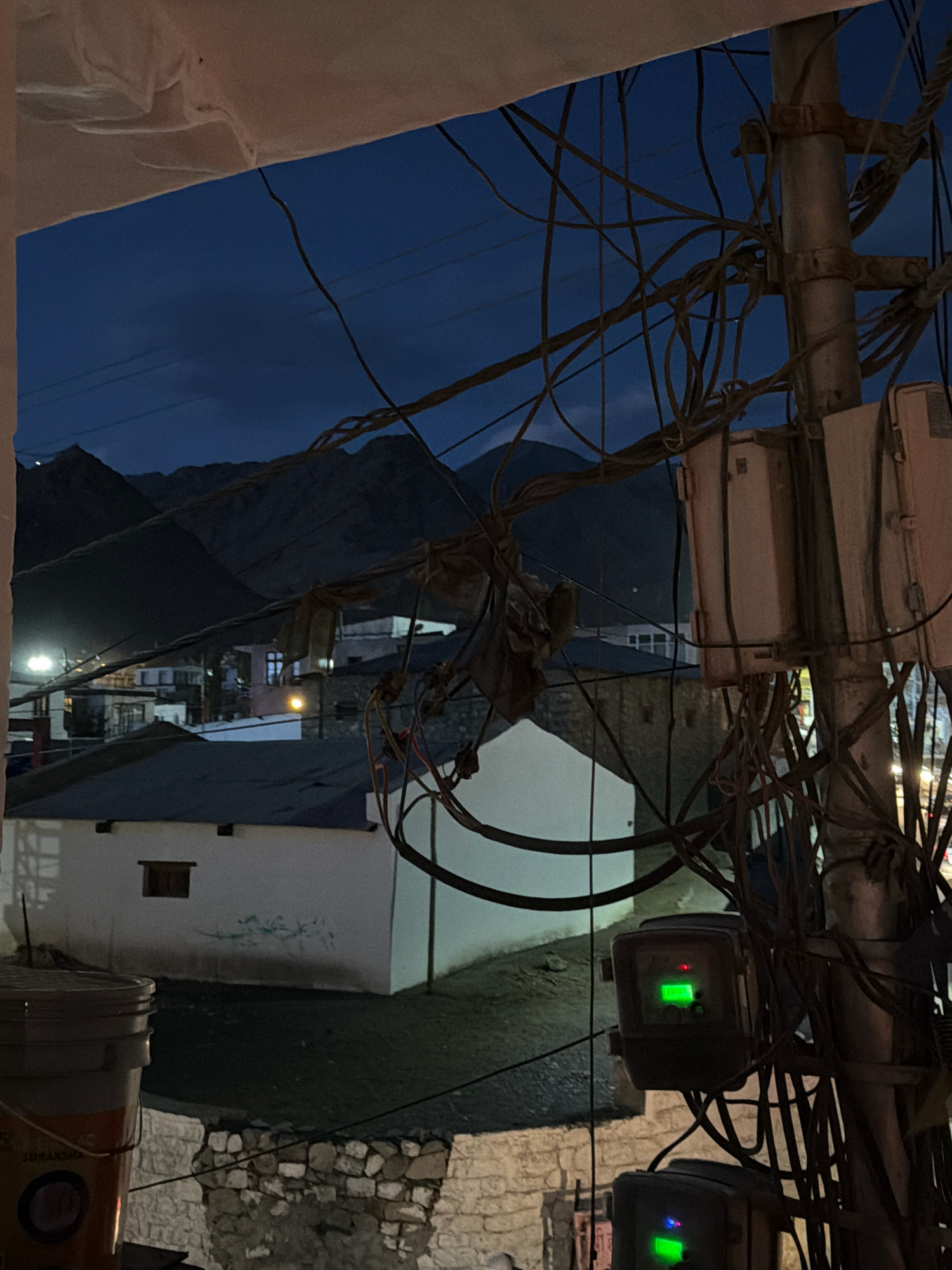 The image captures a nighttime scene in Leh, India, featuring a jumble of electrical wires and utility boxes attached to a pole in the foreground. The sky is a deep blue, suggesting dusk or early night, with silhouetted mountains visible in the background. Several buildings with dark roofs are scattered throughout the landscape, some with illuminated windows. A large white structure with a small dark window is prominent in the mid-ground. A weathered bucket with orange and white detailing is partially visible in the lower left corner. The overall impression is one of a somewhat unkempt but functional urban or semi-urban environment.