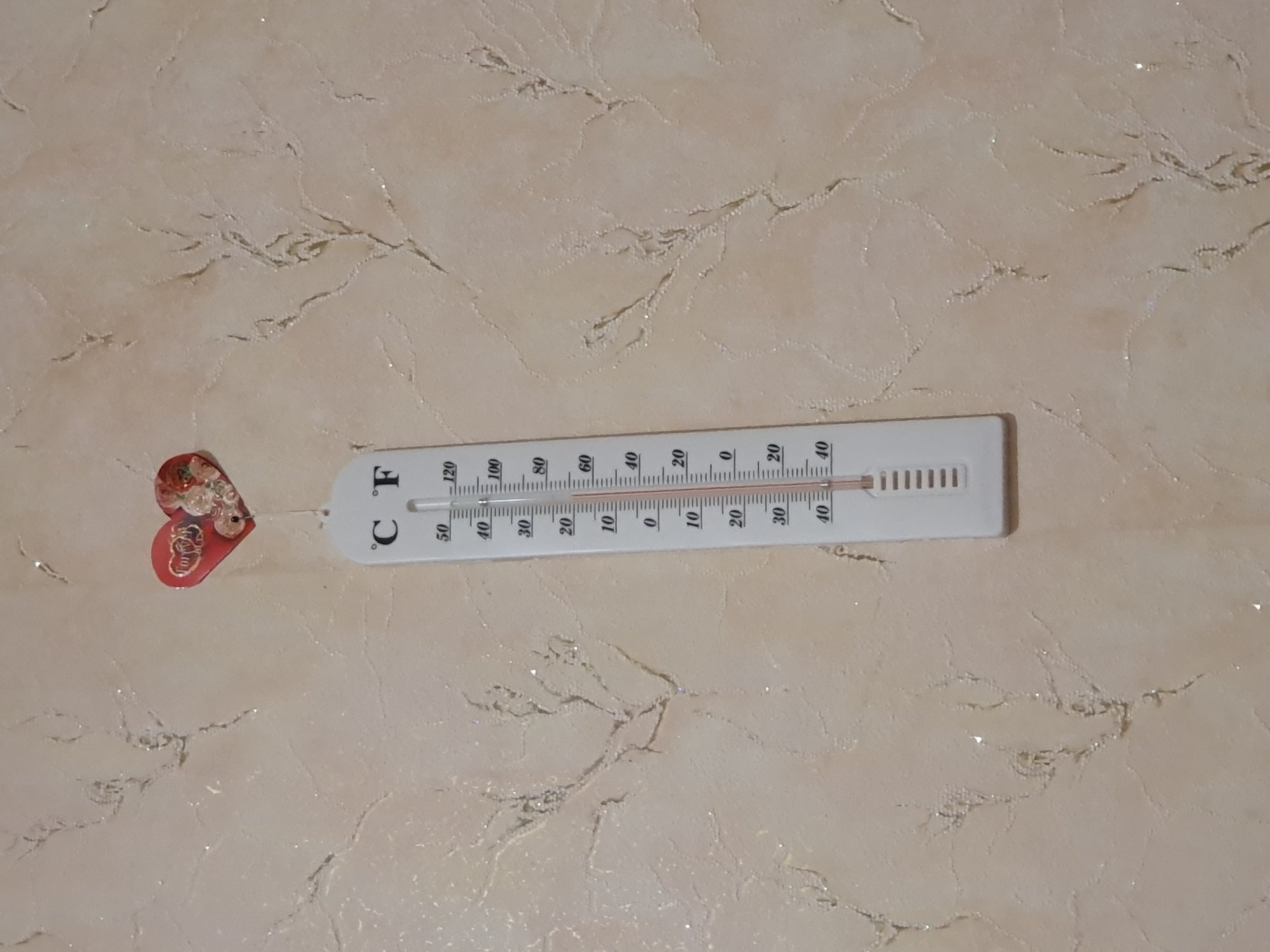 A thermometer hangs on a light-colored wall with a textured pattern. The thermometer is white and has markings for both Celsius and Fahrenheit. The red line indicating temperature is below the 10-degree Celsius mark. Above the thermometer, a small red heart-shaped decoration is attached to the wall. The background wall appears to have a subtle, light-colored pattern resembling branches or vines. The location is not specified in the image or provided data.