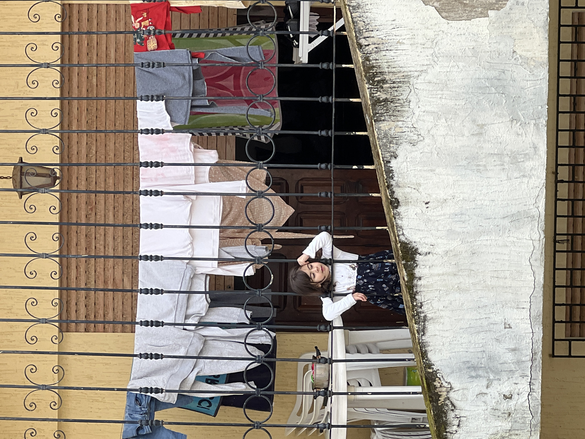 The image, initially rotated, depicts a young girl standing on a balcony in what appears to be a residential setting. She has dark hair and wears a white long-sleeved shirt with a dark, patterned skirt. The girl looks towards the viewer with a gentle smile, her arms resting on a black wrought iron railing adorned with decorative scrolls and rings.
Various items of clothing are hung to dry on a line stretched across the balcony. These include a red t-shirt featuring a reindeer and present design, multiple grey and white t-shirts, a patterned blanket or towel, and blue jeans. A turquoise bag with the partially visible text "CI" is also hanging on the line.
The background reveals a light-yellow painted wall and brown wooden paneling, likely a door or part of the building's facade. Through the railing, a darker interior space is visible, containing several stacked white plastic chairs. To the right, a large, weathered white-painted pillar or wall shows significant wear, with peeling paint, cracks, and patches of dark moss or grime. A grid-patterned element, possibly a window or vent, is partially visible on the far right. A vintage-style metal lantern hangs from an overhead structure on the left side of the balcony. The bright lighting indicates it is daytime, capturing a slice of daily life.