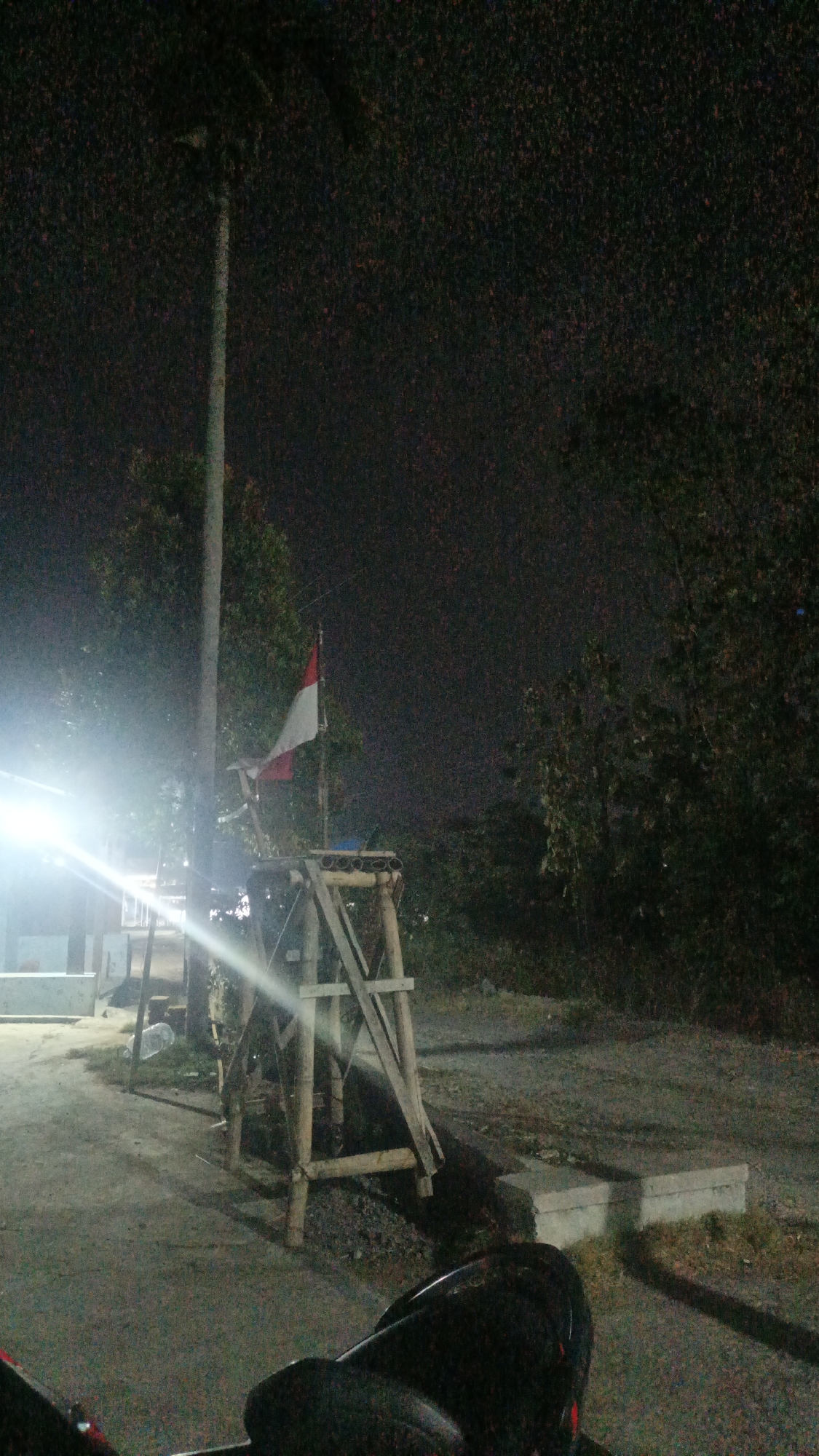 The image depicts an outdoor scene at night, likely in Sambi, Indonesia, based on the context provided. The foreground shows the dark silhouette of a motorcycle, with a helmet visible. The middle ground features a wooden structure supporting a red and white flag, appearing to be the Indonesian flag. The flag is partially illuminated by a bright light source to the left, creating a strong glare that washes out some details. Behind the flag, trees and foliage are visible, their forms softened by the darkness. The ground is a mix of dirt and gravel. The background is predominantly dark, with a dimly lit sky suggesting it is nighttime. A tall pole, possibly a light pole or tree, is also visible to the left of the flag.
