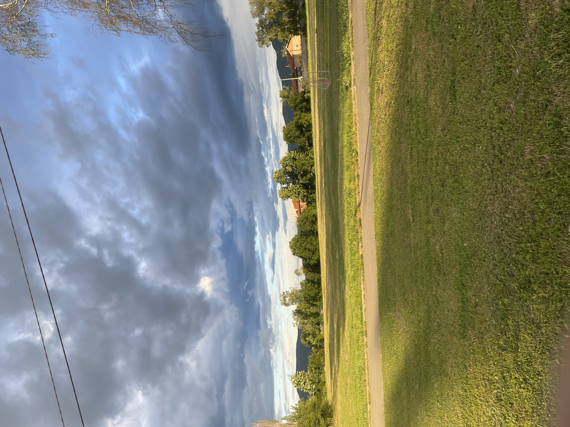 This image captures an outdoor landscape, likely in a rural or semi-rural setting near Ihtiman, Bulgaria. The scene is dominated by a dramatic sky and a verdant expanse of land.
The top portion of the image features a dynamic sky, characterized by heavy, dark clouds on the left, gradually transitioning to lighter, brighter clouds and patches of blue sky towards the right. Some bare tree branches are visible in the upper left corner, along with utility wires extending vertically.
Below the horizon, a continuous line of diverse trees, ranging from dense green foliage to lighter, sparse canopies, stretches across the midground. Behind these trees, distant mountains rise, appearing hazy and soft against the sky. Scattered among the trees and in the distance, a few buildings with reddish-brown roofs are discernible.
The lower half of the image is covered by a wide, well-maintained green grass field, suggesting an open park or pasture. A paved path, possibly concrete or asphalt, runs along the right edge of the field, creating a clear demarcation. The lighting on the grass is uneven, with brighter, sunlit areas contrasting with shaded patches, indicating the sun breaking through the clouds.
The image depicts a tranquil, static scene with no discernible human or animal activity. The weather appears to be partly cloudy, with sunlight illuminating parts of the landscape. The time of day is likely late afternoon or early morning, given the quality of light and shadows. There is no visible text in the image.