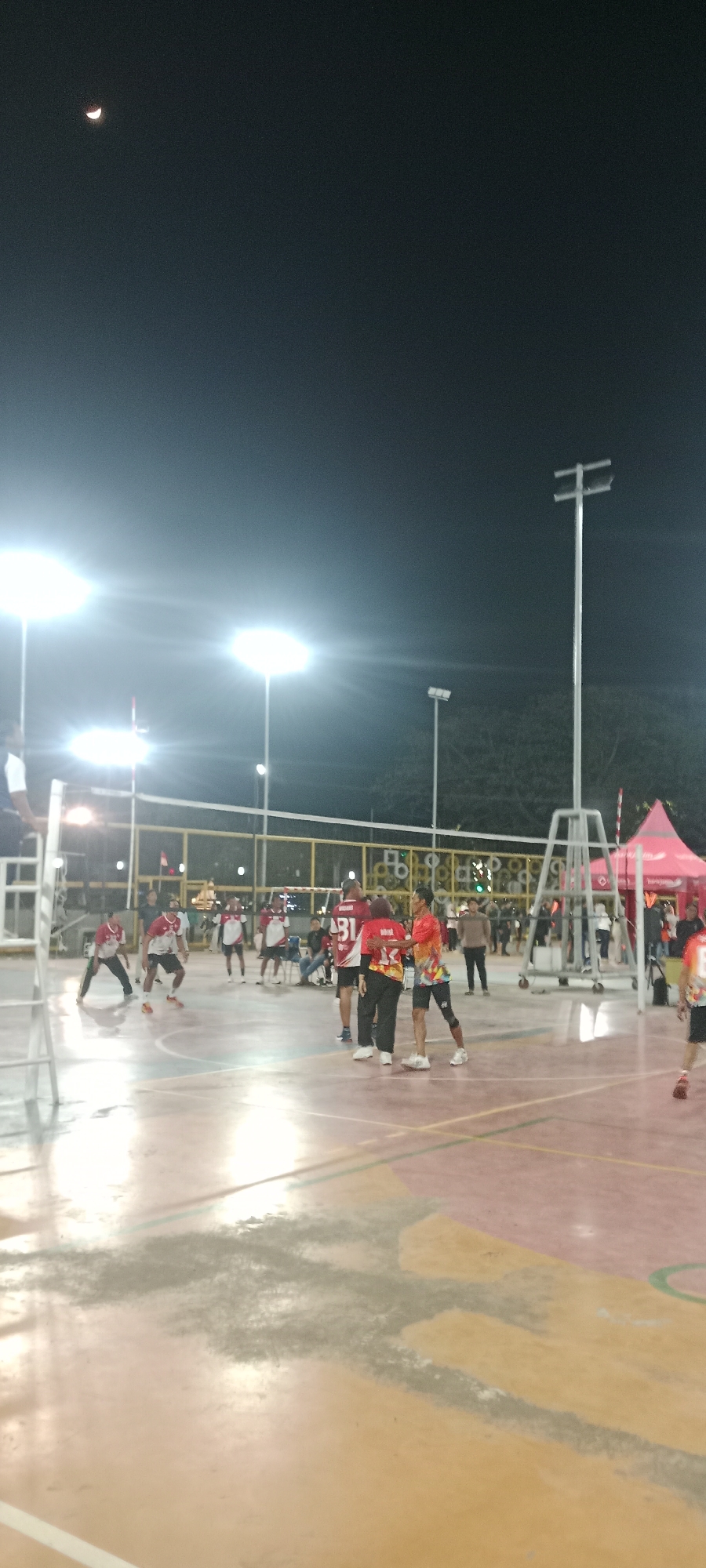 The image captures a nighttime volleyball game in Tuban, Indonesia. Several individuals, dressed in athletic wear, are engaged in the game on an outdoor court illuminated by bright floodlights. The court surface appears to be concrete or asphalt, with visible court markings. In the background, a volleyball net is strung across the court, with structures supporting it. Other individuals are visible in the background, some spectating. The sky above is dark, with a sliver of the moon visible. A pink tent-like structure is also present in the background, possibly indicating a vendor or spectator area. The overall scene suggests a casual, community-level sporting event taking place in an urban or semi-urban outdoor setting in Tuban.
