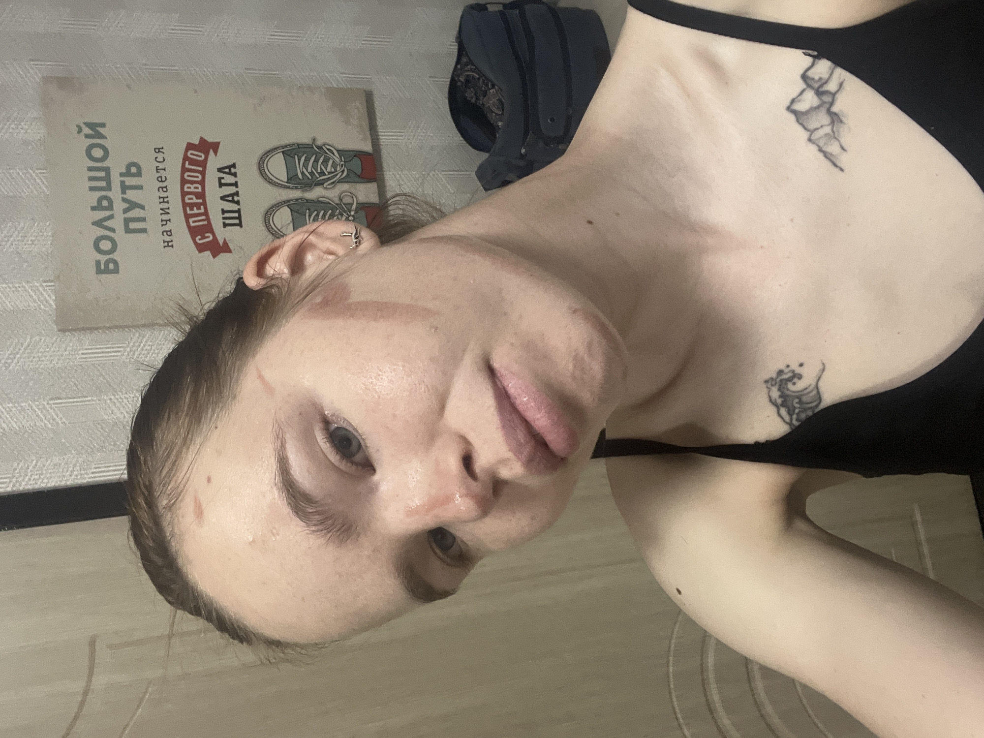 A photograph shows a woman indoors. She is wearing a black tank top and has tattoos on her chest and collarbone. There is a sign on the wall written in Russian that includes text and images of sneakers. A backpack is visible in the background. The location is Perm, Russia.