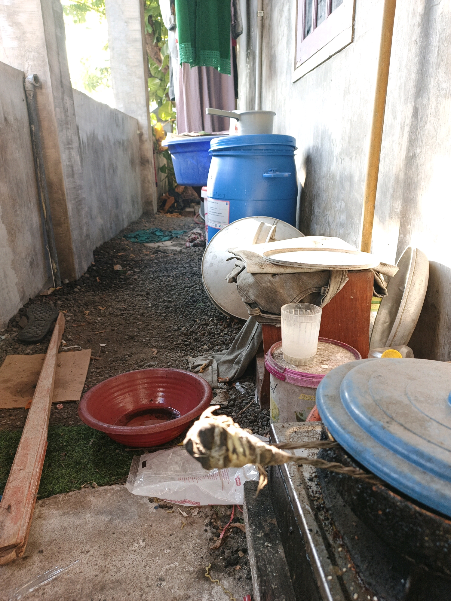 The image depicts an outdoor scene with a narrow alleyway between two textured concrete walls. The ground is covered with gravel and dirt, with patches of green artificial turf visible in the lower left. Various objects are scattered throughout the space, suggesting a storage or work area.
Prominently featured is a large blue plastic barrel, likely a water container, with a white lid. Next to it, a smaller blue basin sits on the ground. A wooden stand supports a white lid and a stack of folded grey fabric, possibly clothing. A clear glass with a cloudy liquid stands on a pink-rimmed bucket.
In the foreground, a red basin filled with dark liquid is placed on the ground. To the right, a piece of twisted rope or wire is visible, attached to what appears to be a metal structure. A small, deceased creature, possibly a bird or reptile, with visible legs and feathers, lies on the rope. A blue plastic lid is positioned in the lower right corner.
On the left side of the image, a worn wooden plank leans against the wall, and a single black sandal rests on the ground. Various plastic bags and debris are scattered on the ground. The scene is illuminated by bright sunlight coming from the left, casting shadows and highlighting the textures of the environment and objects. The overall impression is of a utilitarian outdoor space in a residential or semi-industrial area. The location is specified as Ketegan, Indonesia.