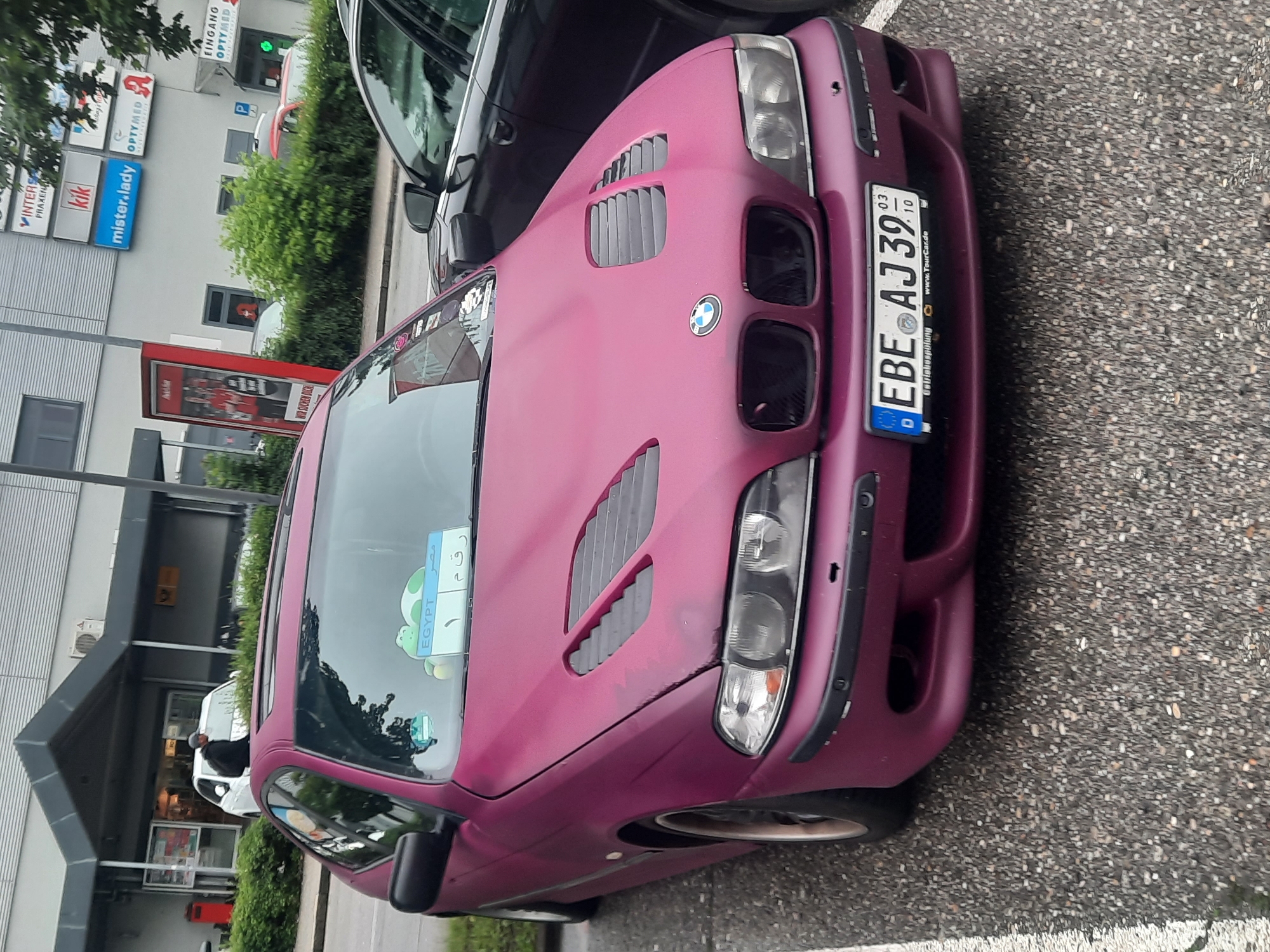 A matte purple BMW 5 Series sedan is parked in a lot. The car has custom hood vents and is seen from a slightly low angle, emphasizing the front of the vehicle. A license plate that reads "EBE AJ39 03 10" is visible on the front bumper. In the background, a building with various signs, including "kik" and "mister.lady," suggests a commercial area. Other cars are parked nearby, and greenery can be seen along the building's facade.