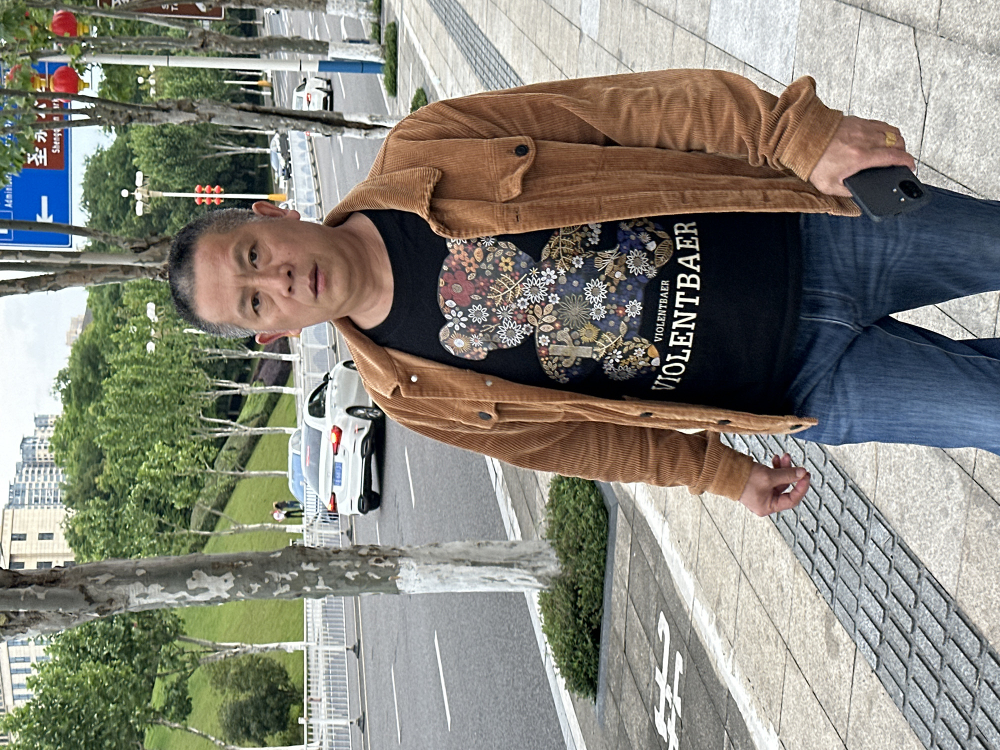 A middle-aged Asian man stands on a sidewalk in what appears to be Jiangjin, China. He is wearing a brown corduroy jacket, a black t-shirt with a floral bear design and the word "VIOLENTBAER" on it, and blue jeans. He holds a black cell phone in his right hand, and his attention is focused on the camera.
The sidewalk is made of light gray stone tiles, and the environment includes trees, grass, a street, and buildings in the background. A white car is visible on the street. Street signs in Chinese are also visible.