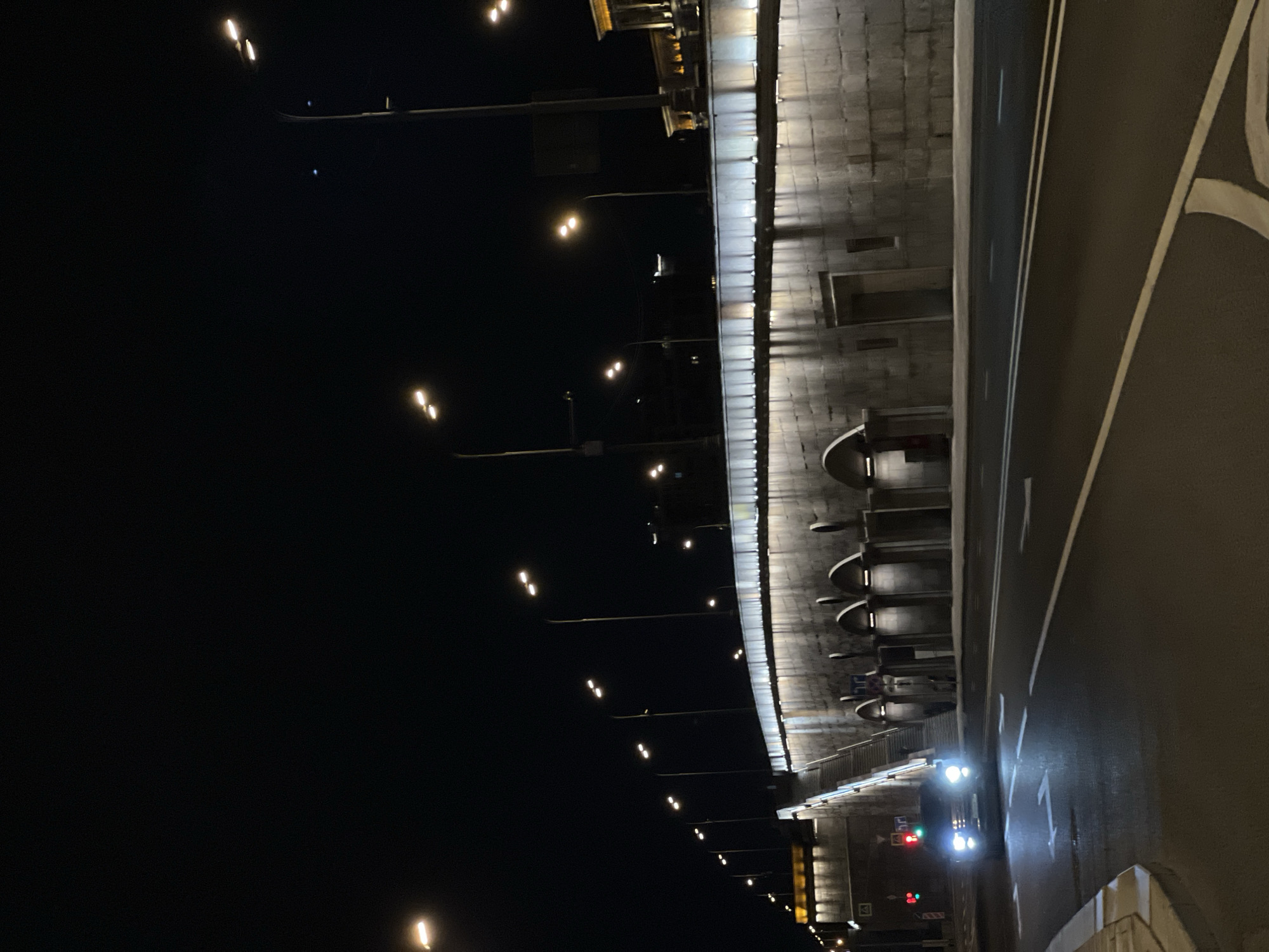 The image depicts a night scene in Moscow, Russia, specifically inside a tunnel. The tunnel structure features gray concrete walls with arched recesses and evenly spaced lighting fixtures along the upper edge. A multi-lane road with painted lane markings runs through the tunnel. A vehicle with illuminated headlights and taillights is visible at the tunnel's end. Street lights are visible outside the tunnel.