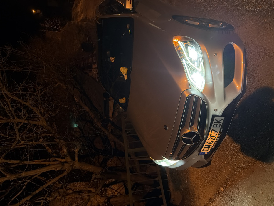 The image captures a silver-colored Mercedes-Benz sedan parked at night in Podvis, Bulgaria. The vehicle is angled towards the viewer, with its headlights illuminated, projecting bright white and amber light. The front bumper displays a Bulgarian license plate, "CM 6207 BK." On the dashboard, visible through the windshield, are two yellow, duck-shaped figures and a small, dark object suspended near the rearview mirror. The car features a dark-colored roof.
In the background, to the left of the vehicle, stands a large, leafless deciduous tree with a dense network of bare branches. A light-colored metal fence or railing runs horizontally between the car and the tree line. The ground beneath the car is dark and appears to be an uneven, possibly paved or gravel, surface. In the upper right background, a distant street light emits a faint orange glow. A subtle, diffuse green light source is barely discernible through the tree branches on the left side of the frame. No individuals are present.