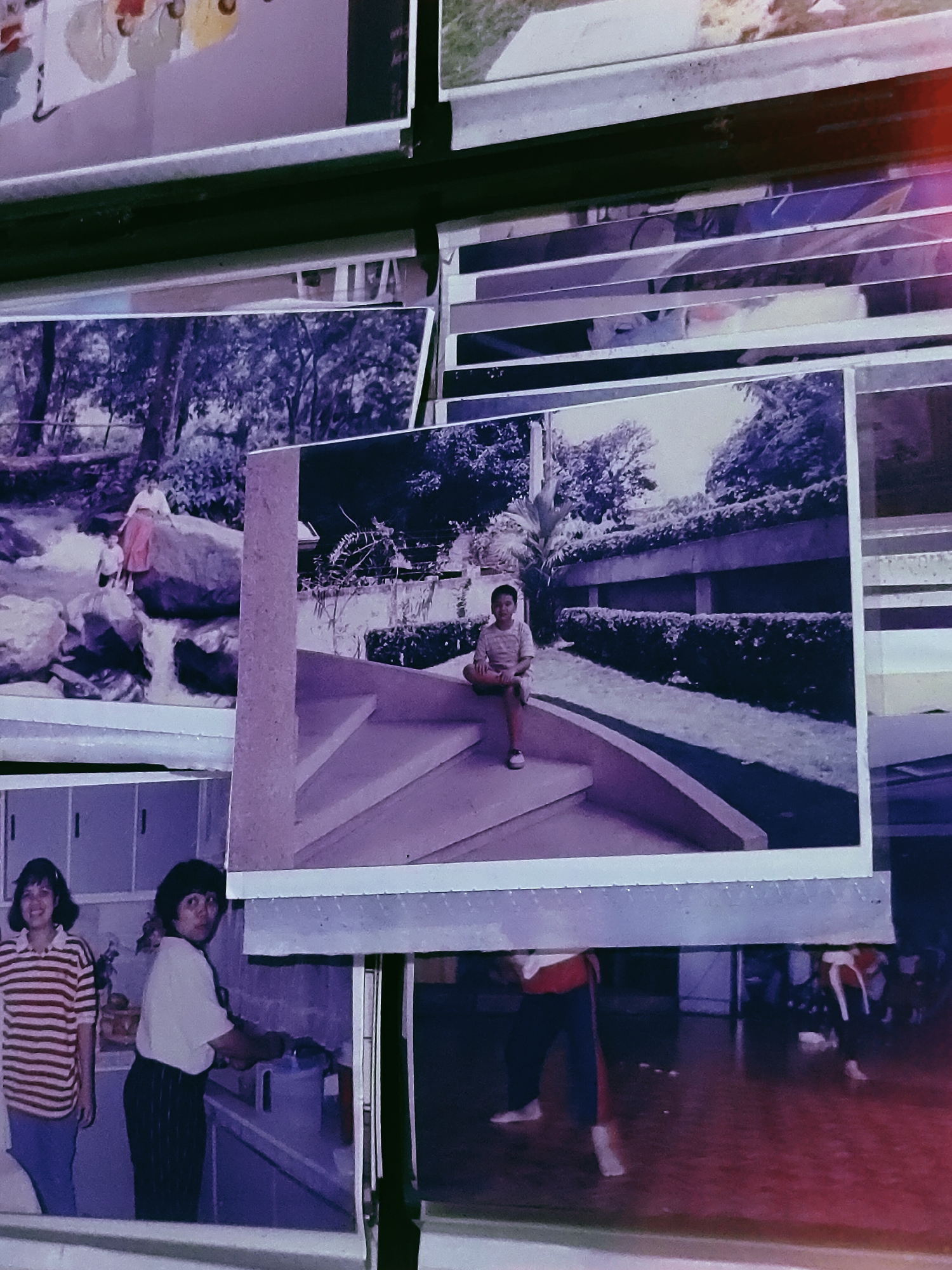 The image displays several aged physical photographs, some overlapping, presenting a compilation of past events. The dominant color cast across all prints is purple and desaturated.
In the central, prominent photograph, a young boy is seated on curved concrete steps outdoors in Davao City, Philippines. He is wearing a light-colored short-sleeved shirt and shorts, with white sneakers visible on his left foot. Behind him, a landscaped area features a long row of trimmed hedges, a concrete retaining wall, and numerous tropical trees and dense foliage under an overcast sky.
To the left of the central photo, another image depicts a woman in a light top and long skirt, accompanied by a small child, standing on large rocks alongside a small waterfall or stream. This scene, likely within a natural park in Davao City, is surrounded by lush trees and undergrowth.
In the bottom left, a photograph shows two women in what appears to be a kitchen or utility room within Davao City. The woman on the left, with short dark hair, is smiling and wearing a red and white striped t-shirt and blue pants. The woman on the right, with short dark curly hair, is looking towards the camera while tending to items on a counter. White cabinets are mounted on the wall behind them.
A partially visible photograph at the bottom right reveals two individuals, likely engaged in physical training such as martial arts or dance, within a large indoor space with a reddish-brown floor in Davao City. They are barefoot and in motion, wearing dark uniforms with contrasting red and white accents.
At the top, a fragment of an abstract artwork or painting with light colors and shapes is partially visible. The overall scene suggests a collection of personal memories captured on film.