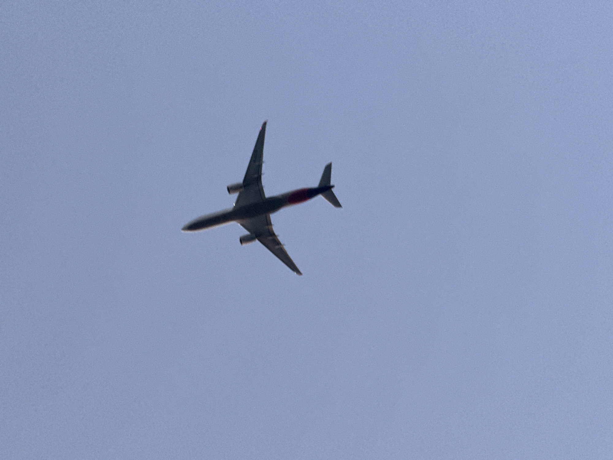 An airplane is visible flying across a clear blue sky. The underside of the airplane is visible, and it appears to be a passenger jet with its wings extended and engines attached. The airplane is angled slightly upwards, suggesting it is either taking off or climbing. The image does not provide any specific details about the location in Castelldefels, Spain, other than that an airplane is flying overhead.
