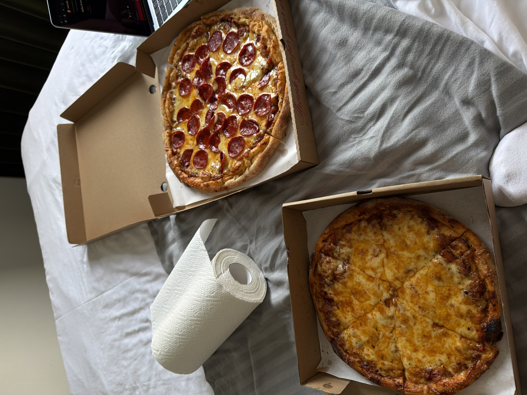 The scene shows two pizzas in open cardboard boxes on a gray and white striped bed. One pizza has pepperoni toppings, while the other appears to be a cheese pizza. A roll of paper towels lies beside the pizza boxes. A laptop is partially visible in the upper left corner of the frame. The environment is casual and domestic. There is no information in the image to suggest it is in Đà Nẵng, Vietnam.