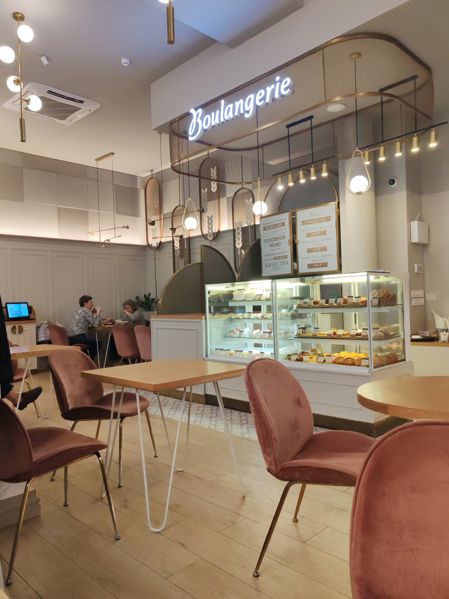 The image displays the chic interior of a "Boulangerie" (bakery/cafe), designed with a modern and elegant aesthetic, likely located in Saint Petersburg, Russia, given the Cyrillic text on its menus. The atmosphere appears bright and inviting, illuminated by a combination of ceiling spotlights, decorative gold-toned chandeliers with spherical bulbs, and track lighting. An air conditioning unit and a security camera are also visible on the ceiling.
The decor includes light wooden flooring, white wainscoting on the lower walls, and a subtle geometric pattern on the upper sections. Gold accents are prevalent throughout, featured in the light fixtures and an elegant arched metal framework above the main counter. Seating comprises small, light-wood tables with either white hairpin or gold-finished metal legs, complemented by comfortable dusty rose velvet upholstered chairs.
In the background, two individuals are seated at a table, engaged in conversation, contributing to a relaxed ambiance. The central feature of the serving area is a brightly lit glass display case, showcasing a variety of baked goods such as croissants, rolls, cakes, and pastries. Above this, a glowing neon sign prominently spells out "Boulangerie."
Two framed menus, written in Russian, are displayed behind the counter. The left menu advertises "Breakfasts all day long," listing "Syrniki, Porridges, Omelette," and highlights a "MAIN MENU" focused on "natural products of HIGHEST QUALITY." The right menu details "Lunches" available "From 12:00 TO 17:00," offering meal combinations like "Salad, Soup, Drink" for 470 P, "Salad, Soup, Hot dish" for 560 P, and "Salad, Soup, Hot dish, Drink" for 660 P (Rubles). The scene depicts customers enjoying the offerings of this stylish cafe.