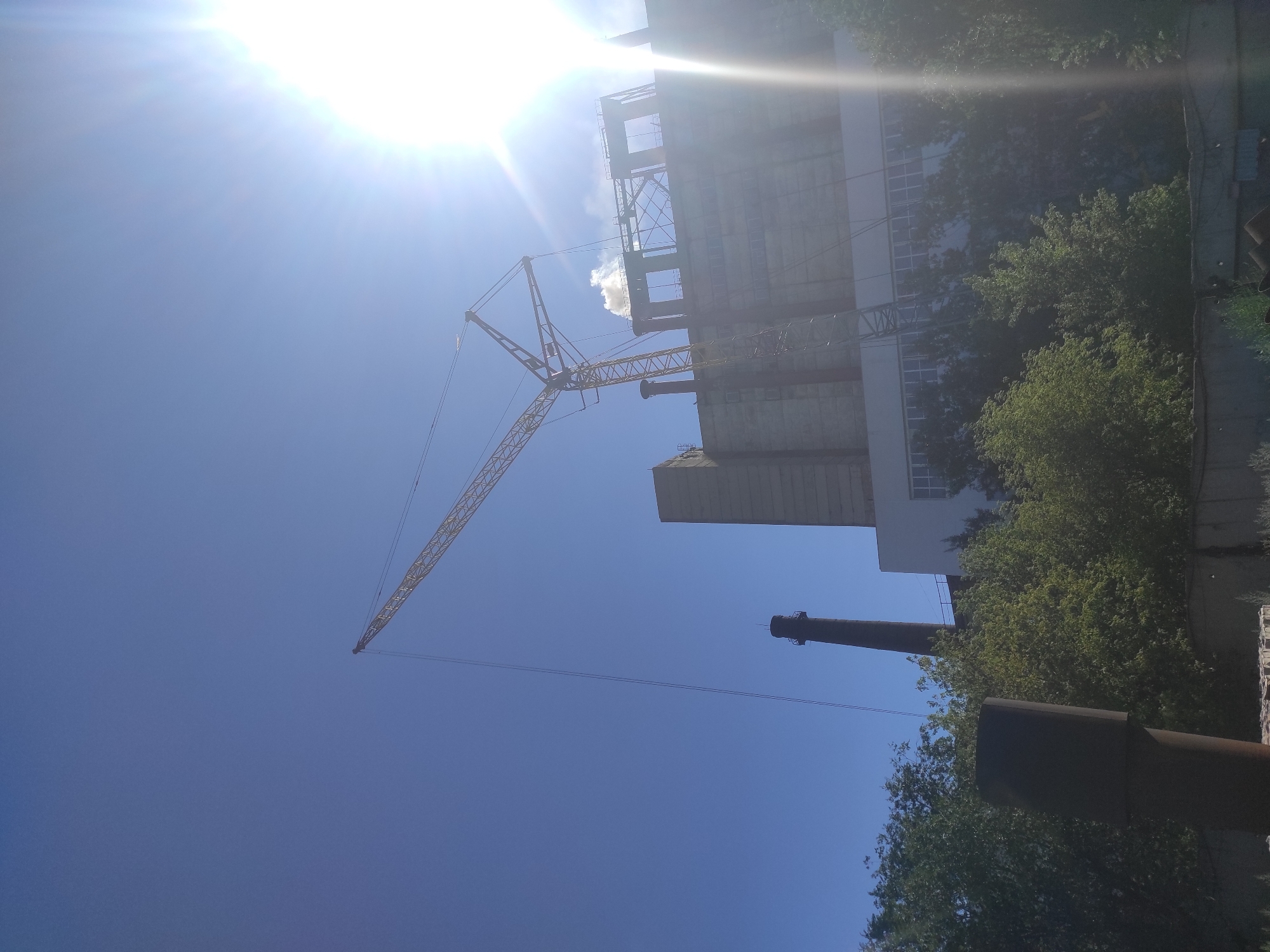 The image shows a large industrial facility with a tall chimney and a construction crane against a clear blue sky. A bright sun is visible in the upper right corner, causing lens flare. The building appears to be made of concrete and has large windows. There is some steam or smoke rising from the top of the building. The foreground shows trees and a dark, cylindrical structure. The overall scene suggests an active or recently active industrial site, possibly undergoing construction or maintenance. The specific location in the city of Kyzyl, Kazakhstan, is not discernible from the image itself, as it lacks any identifying landmarks or textual information.
