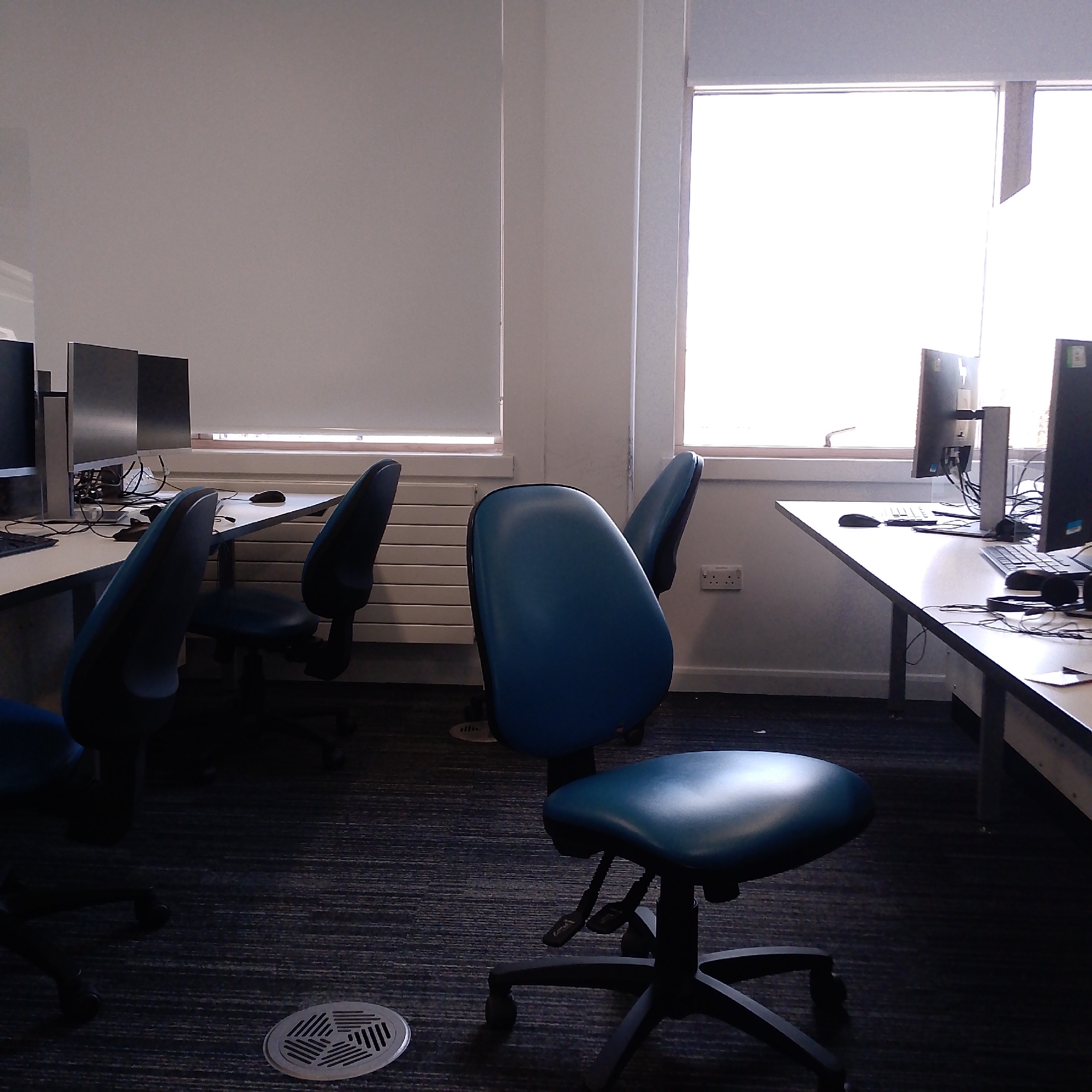 The photograph depicts an interior space, possibly an office or computer lab, in Coventry, United Kingdom. The room contains several computer workstations with flat-screen monitors and keyboards on light-colored desks. Blue ergonomic chairs are positioned at the desks, some of which are unoccupied. There are two windows in the background with closed roller blinds, letting in light. The walls are painted light gray. The floor is carpeted with a dark gray patterned rug. A circular air vent is visible on the floor near the center of the frame. There are electrical outlets on the wall beneath the window on the right. There is no depiction of individuals present.
