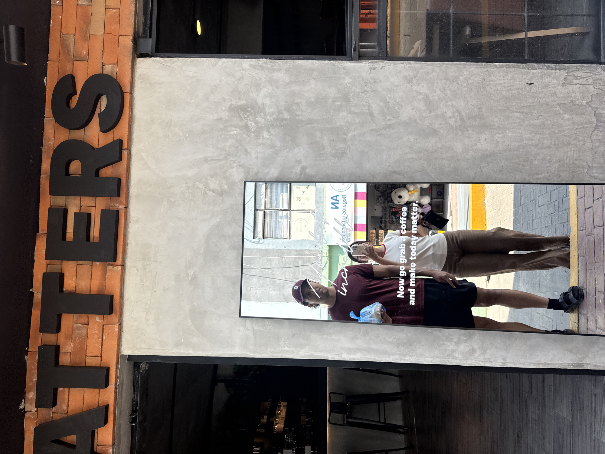 Here is a detailed description of the image:
**Scene**: This photograph shows the exterior of a cafe named "ATTERS" in San Andrés Cholula, Mexico.
**Environment**: The facade combines brickwork with a smooth, gray concrete-like wall. A dark-framed window is positioned above the concrete portion. A modern, sleek sign with the cafe name is mounted vertically on the brick section.
**People**: The photograph is not of people directly, but a reflection of a pair of people captured on a video that shows on a tall monitor attached to the building. One person wears a maroon shirt, a dark baseball cap, and black shorts. The other person is wearing a white t-shirt and brown pants.
**Objects**: The monitor appears to display a video with text overlaid. The text reads "Now go grab a coffee and make today matter" along with other text that is partially illegible. Other objects include chairs and a bar inside the cafe that is visible behind the monitor.
**Actions**: The video seems to show the people casually walking.