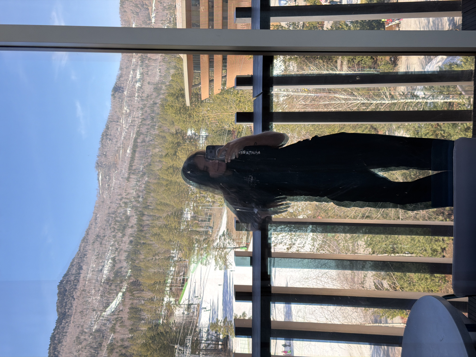 A reflection in a window captures a woman holding a phone while standing on a balcony with a view of mountains and a ski resort. The reflection includes part of the balcony's structure, featuring horizontal slats. The backdrop is dominated by a mountain covered with trees and patches of snow, under a clear blue sky. Ski slopes are visible below the mountain. The exact location in Озёрное, Russia cannot be determined from the image.