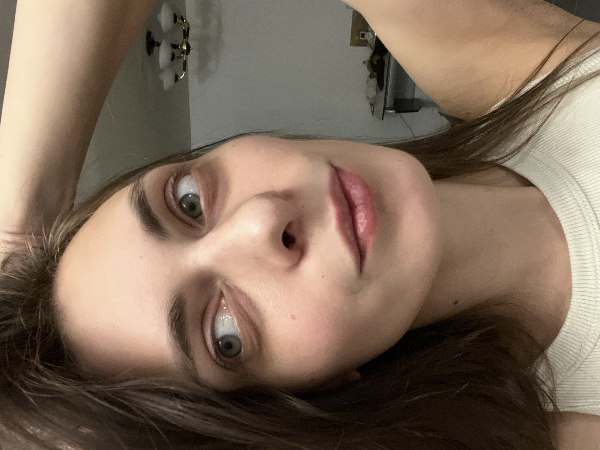 This close-up, angled portrait captures a woman indoors in Krasnoyarsk, Russia. The image is framed as if taken from an unusual perspective, possibly a selfie with the camera held above her, or while she is lying down. The woman has fair skin, long dark hair, and captivating greenish-grey eyes, which are wide and look directly into the camera. Her expression is neutral but direct, with slightly parted, rosy lips. She appears to be wearing a light-colored, possibly white or off-white, ribbed top or tank top. Her right arm is visible in the upper portion of the frame, partially obscuring the background.
The background shows a plain, light-colored wall, on which a decorative wall sconce with two white, bell-shaped lampshades is mounted. The lighting is soft and even, consistent with an indoor setting, possibly during the evening or in an area with controlled illumination. There is no visible text in the image. The overall impression is an intimate, candid moment.