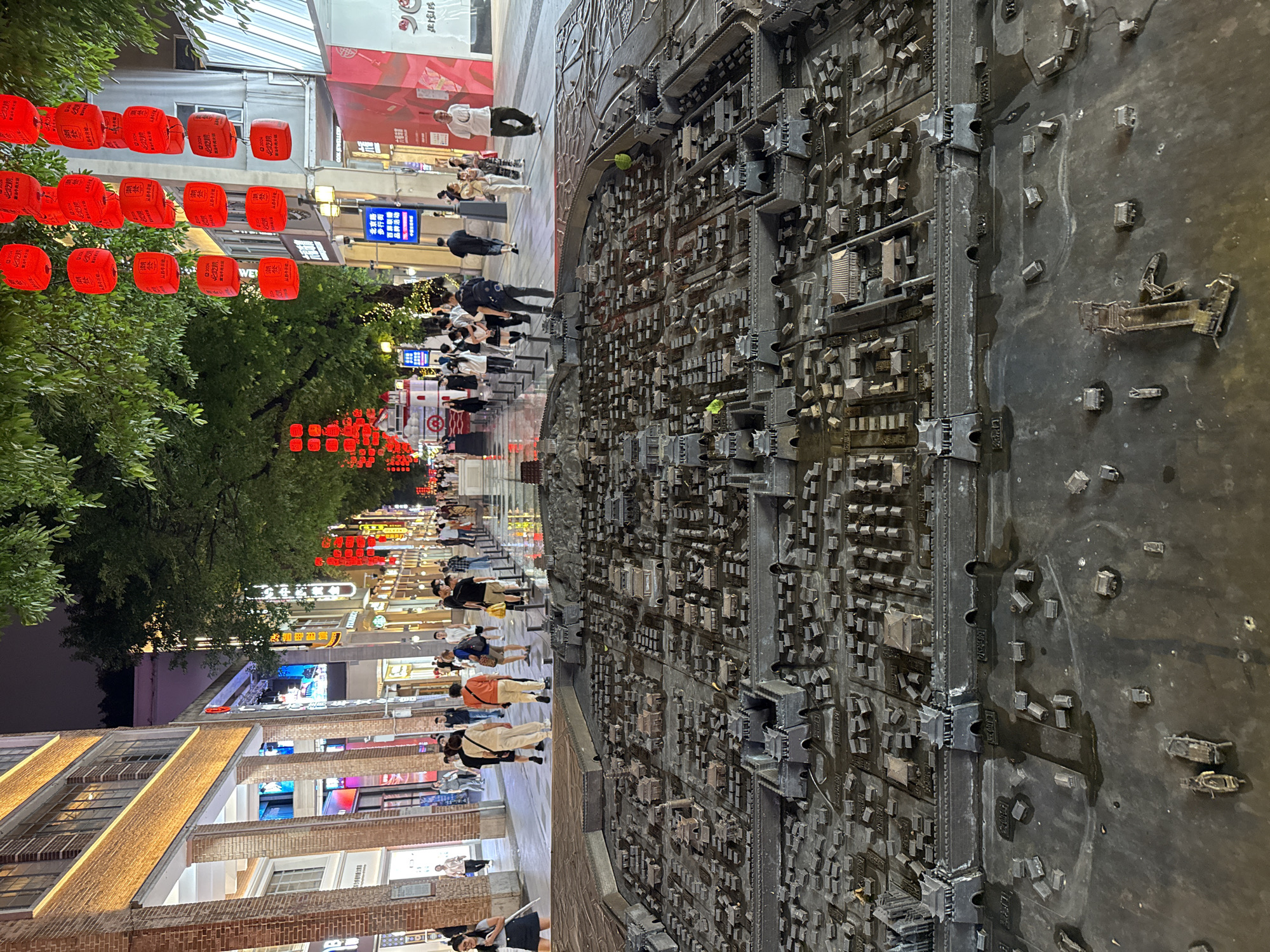 The image shows a pedestrian street in the Yuexiu District of Guangzhou, China at night. Red lanterns hang from the trees. Pedestrians stroll along the street. On the right side of the frame is a detailed model of the city, depicting buildings and streets. The street is lined with shops and brick buildings with illuminated signs.