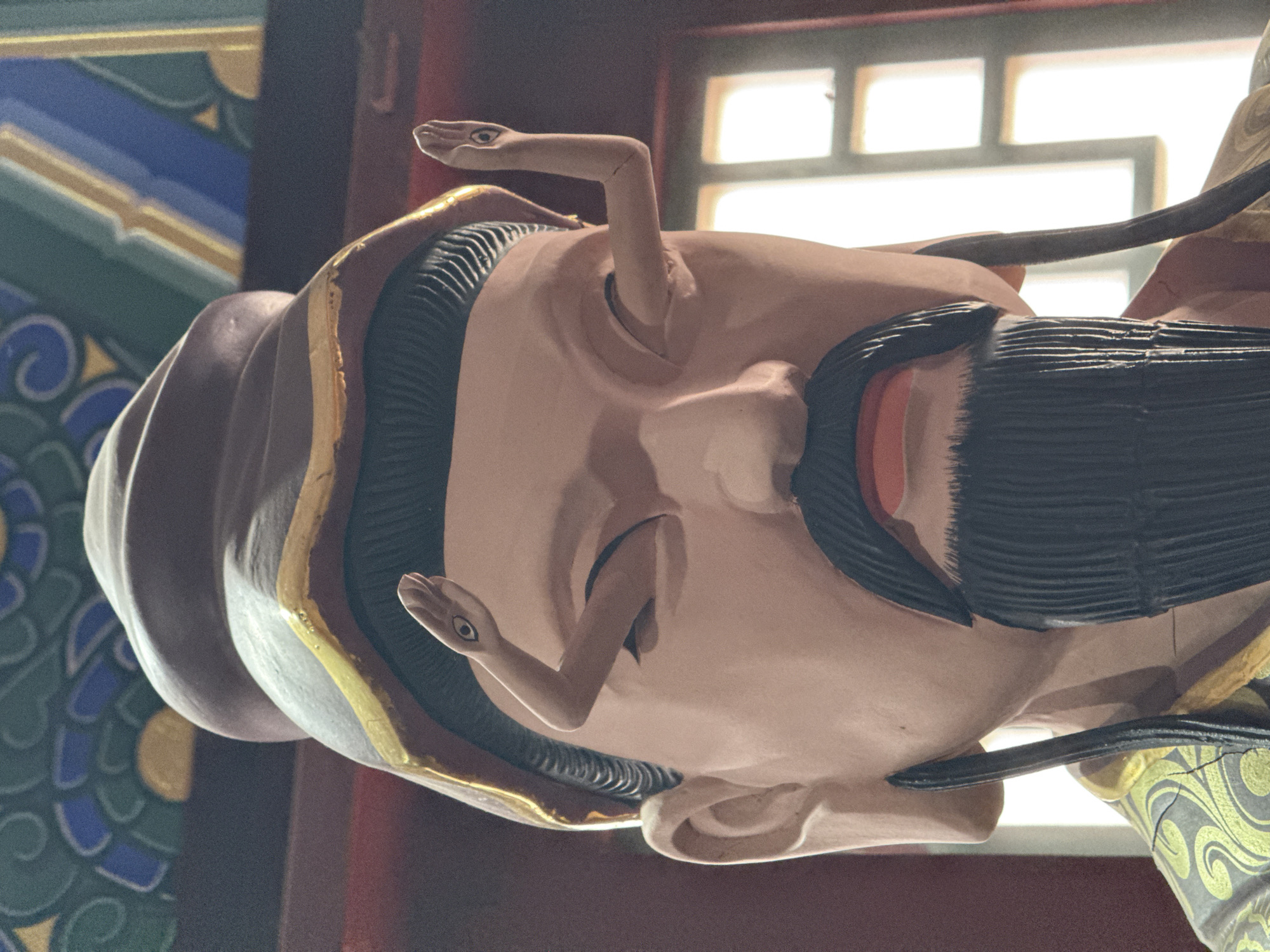 The image shows a close-up of a statue's head. The statue has light skin, a black mustache, and a black hat with gold trim. Two snake heads emerge from the head near the eyes. The background includes a window and a decorative painted ceiling with green, blue, and gold patterns. It's impossible to verify the statue's location in Lintong District, China, based solely on the image.