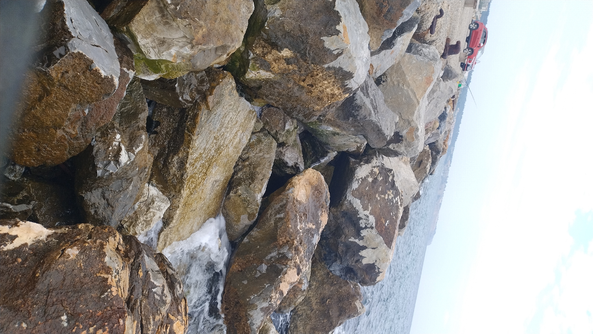 The image captures a rocky coastline in Neoi Epivates, Greece. A red SUV is parked on a paved area to the left, near some rusty metal objects that appear to be mooring equipment. A fishing rod is set up near the car. The foreground is dominated by large, rough rocks, some wet from the sea. The sea is visible in the background, with waves gently lapping against the rocks. The sky is overcast with a few hints of clouds. The overall scene depicts a coastal area with recreational activity.