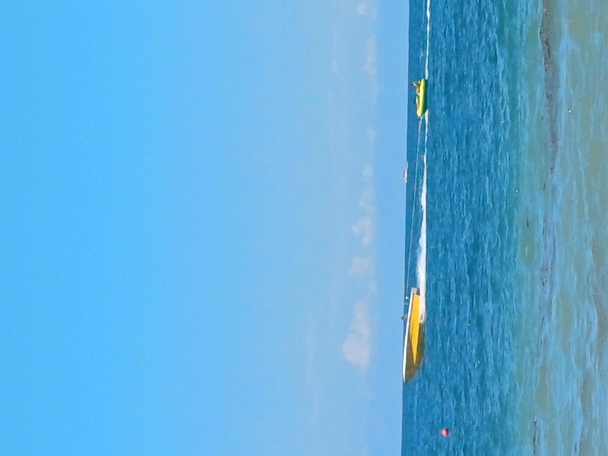A wide-angle, eye-level, outdoor shot captures a bright, sunny day at a beach, likely in Sochi, Russia, given the clear blue sky and azure water. In the middle ground, a yellow and white speedboat is cutting through the waves, leaving a white spray in its wake. A person is visible at the helm of the speedboat. Further out, to the right, a yellow inflatable tube, also known as a "banana boat," is being towed by an unseen vessel, with one person reclining on it. In the far distance, another small white boat is visible. A red buoy floats near the left edge of the frame. The water is a deep blue, with visible ripples and waves. The sky is a clear, vibrant blue with a few scattered white clouds near the horizon.