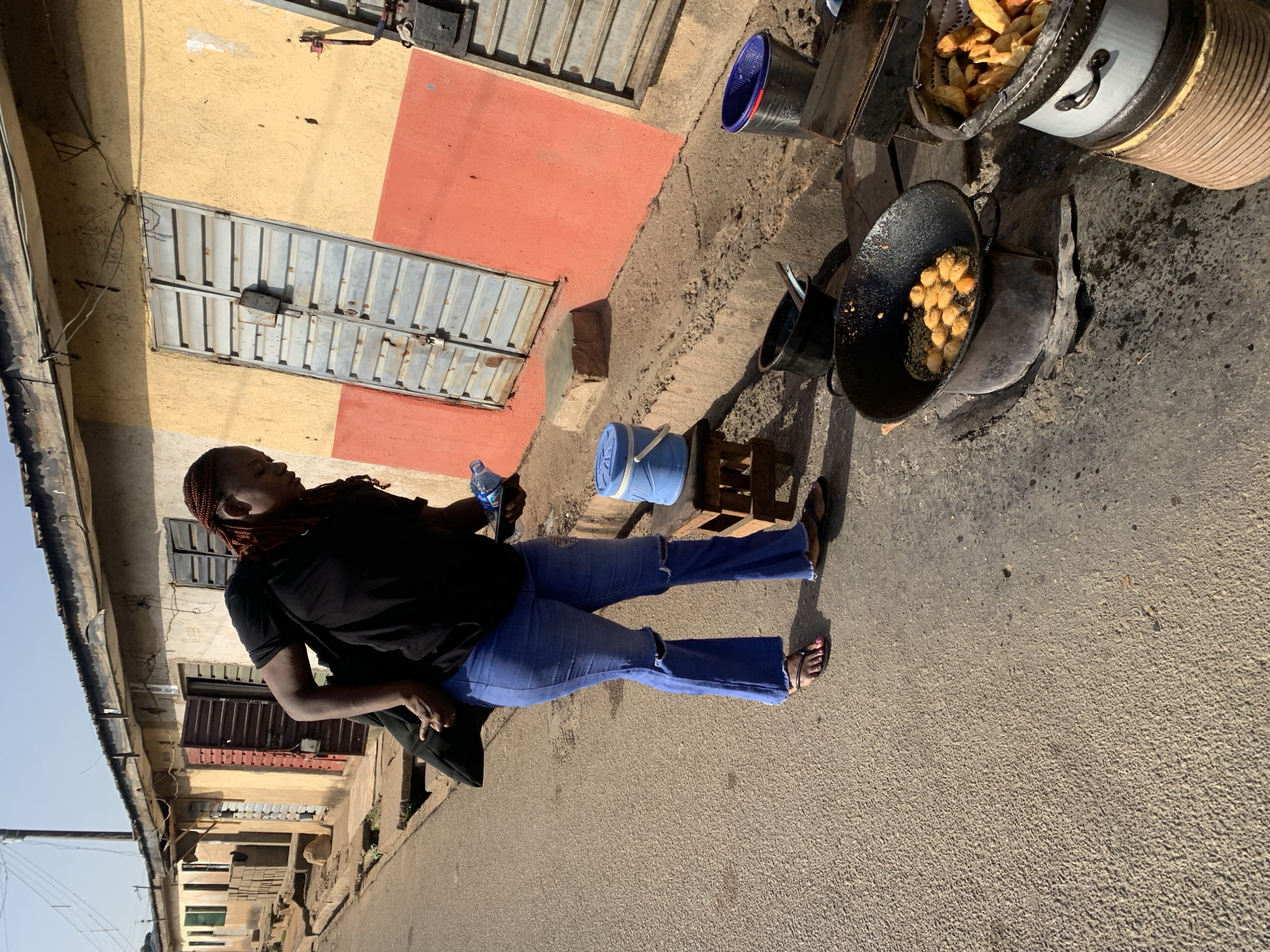 The image depicts a woman standing on a paved street in Jos, Nigeria, near a food preparation area. She is wearing a black t-shirt and blue jeans and is holding a water bottle. To her right is a cooking setup with two large pots and a black pan, likely used for frying food. One pot appears to contain plantain chips, while the other holds fried balls. The background includes buildings with yellow and red walls and corrugated metal doors. A blue bucket sits near a wooden pallet. The street appears to be a public space where food is being sold or prepared.