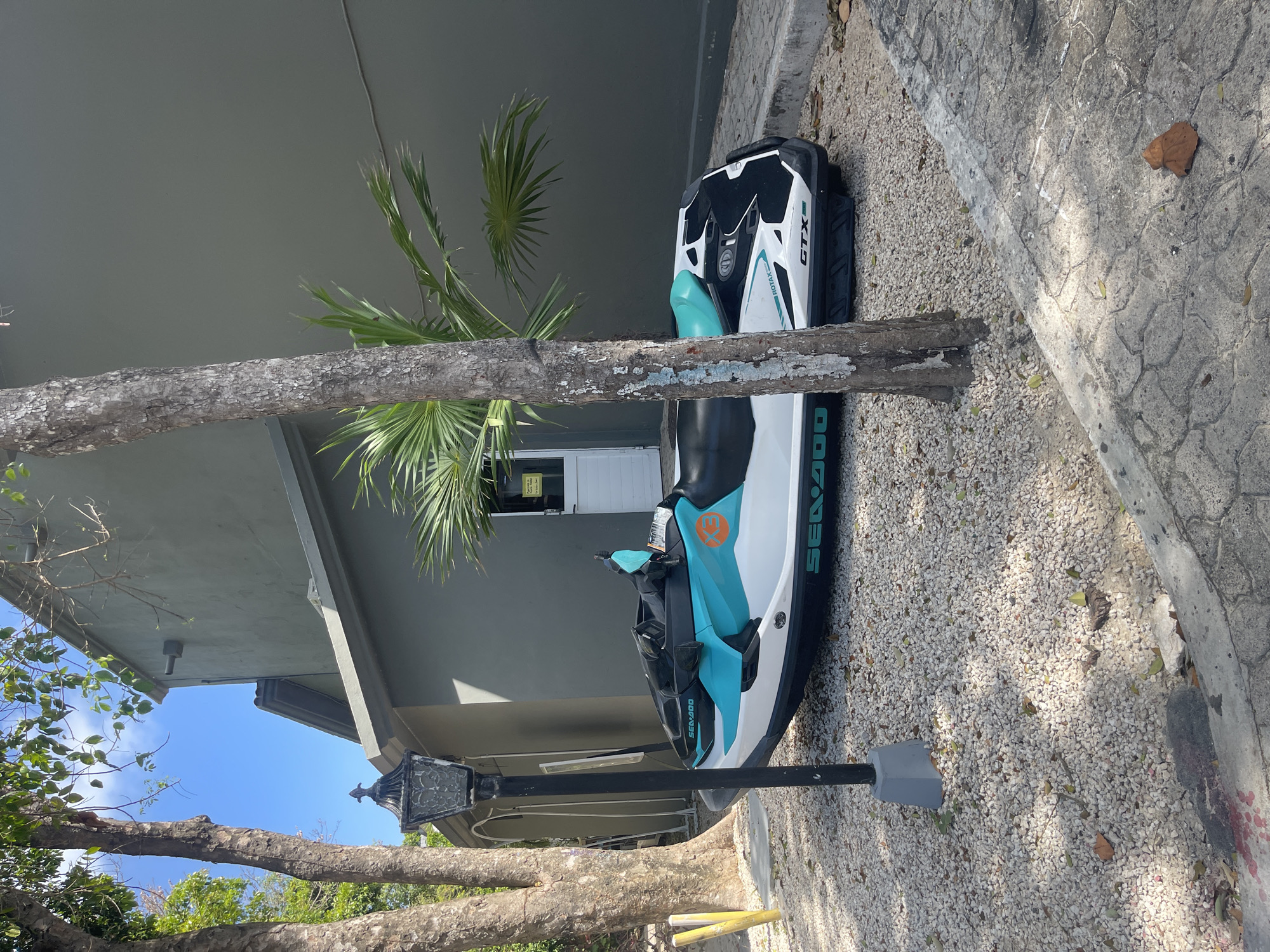 The image shows a white and teal Sea-Doo personal watercraft (PWC) and its black and white GTX cover parked on a gravel area next to a building. A tree trunk and branches obscure parts of the PWC. Behind the PWC, there is a light grey building with a closed white door and a small palm tree. A black lamppost extends upwards near the watercraft. The scene appears to be located outdoors, likely in Playa del Carmen, Mexico. The ground is covered in small white and light grey gravel, bordered by a concrete sidewalk. The weather seems sunny.