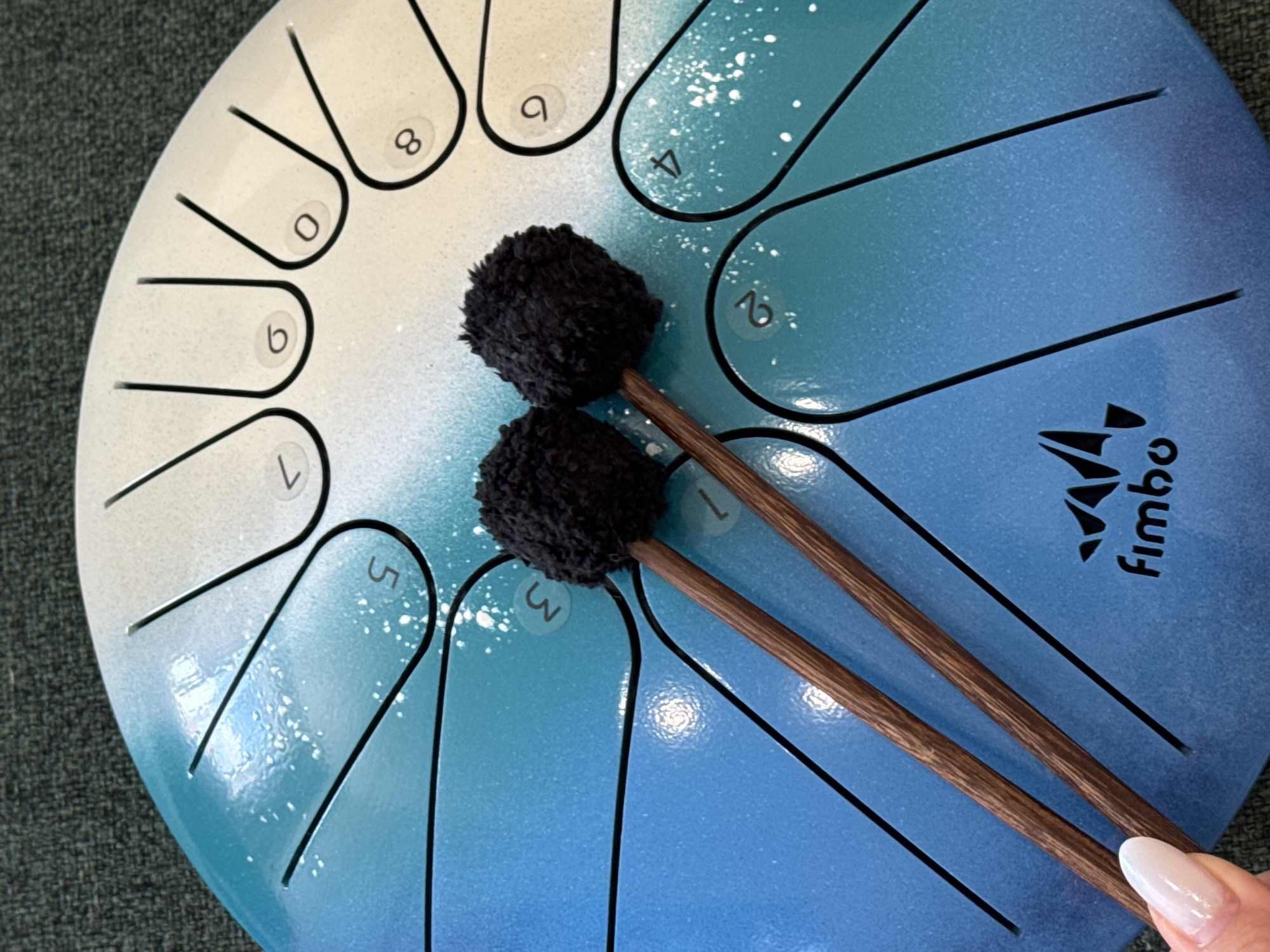 This is a close-up, overhead shot of a blue and white steel tongue drum, with two black mallets resting on its surface. The drum is decorated with a gradient of blue and white, speckled with white paint. Each of the drum's twelve tongues is numbered, with numbers 0 through 9 visible, along with a partially visible '1' and another unreadable number. The mallets have wooden handles and fuzzy black heads. In the bottom left corner, a fingernail with white polish is visible, suggesting a person's hand is holding or interacting with the drum. The background is a dark, textured surface, likely a rug or fabric. The "Fimbo" logo is visible on the drum near the bottom center. The scene is indoors and the lighting is bright and even, illuminating the details of the drum and mallets.