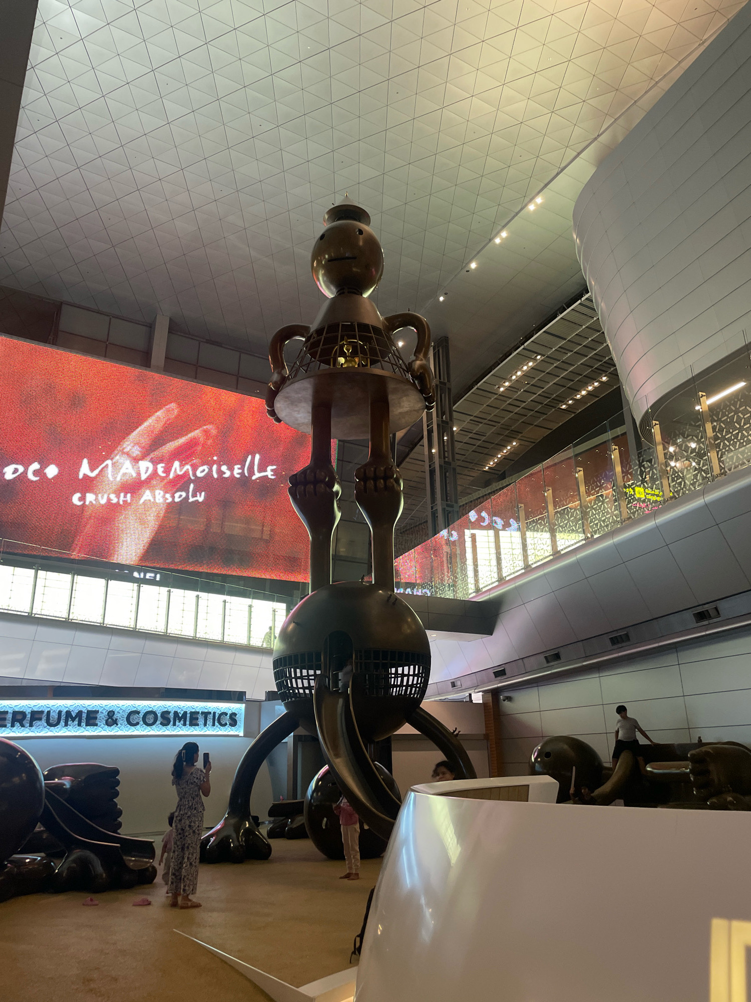 This image captures a wide shot of the interior of Hamad International Airport in Doha, Qatar. Dominating the center of the frame is a large, golden, abstract sculpture resembling a stylized, elongated figure. The sculpture features a spherical head with a simple face, a cage-like structure for its torso, and multiple articulated limbs. The sculpture is positioned on a raised platform, surrounded by other dark, abstract sculptural elements that appear to be part of the same installation.
In the background, a large LED screen displays an advertisement with the text "CO MADEMOISELLE" and "CRUSH ABSOLU," suggesting a luxury brand or perfume promotion. The airport's modern architecture is evident with a high, tessellated ceiling and curved white walls. Several people are visible within the scene. A woman in a patterned jumpsuit stands to the left, holding a phone and seemingly taking a photograph of the sculpture. Another person, possibly a child, is standing near her. Further in the background, a person in a grey t-shirt is visible standing on one of the sculptural elements. A sign reading "PERFUME & COSMETICS" is illuminated in blue and white to the lower left of the image, indicating a retail area. The overall lighting suggests an indoor environment, typical of an international airport terminal.