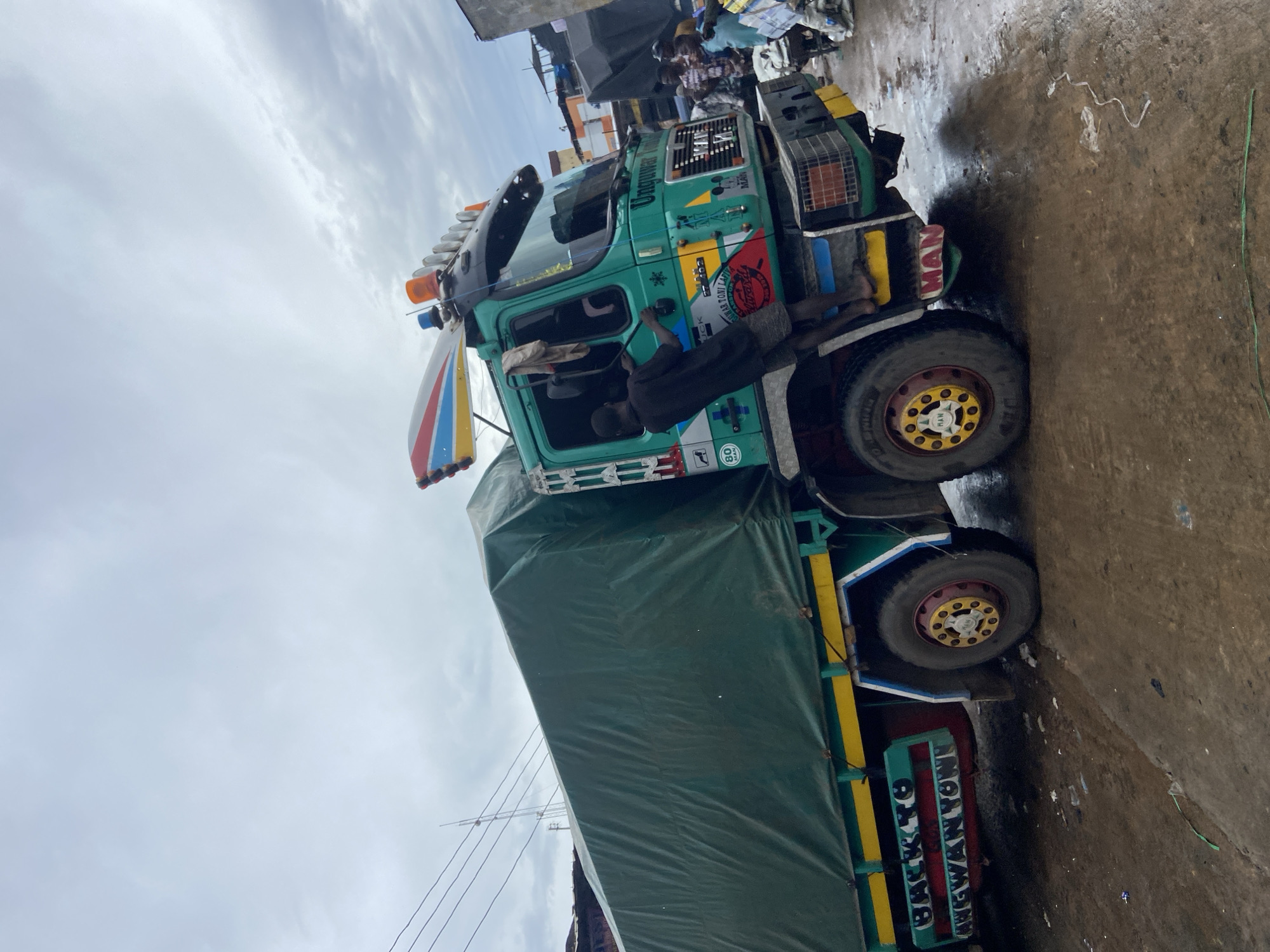 In Ogbunike, Nigeria, a man is seen climbing the side of a green truck that has a large green tarp covering its cargo. The truck has bright yellow, red, and blue accents on its body and wheels. A colorful, wing-like decoration with red, blue, and yellow stripes is attached to the top of the truck's cab, along with several flashing lights. The truck has "Unoguway Transporter" written on the side of the cab. The ground is wet and appears to be made of dirt or mud, with scattered debris. In the background, other trucks and people are visible, suggesting a busy marketplace or loading area. The sky is overcast.