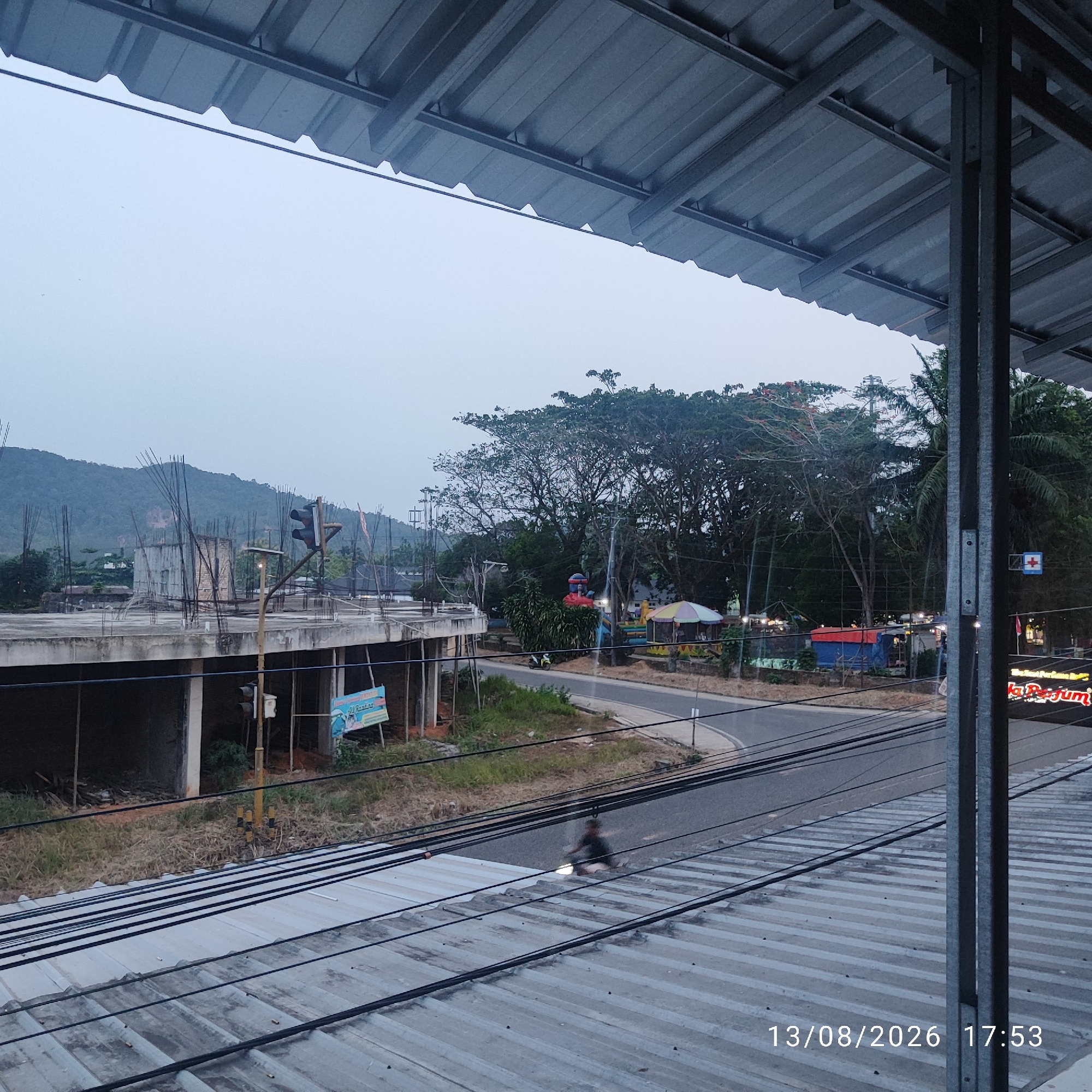The image depicts an outdoor scene under a grey, overcast sky, likely in Buding, Indonesia, as suggested by the watermark "13/08/2026 17:53". The foreground is dominated by a corrugated metal roof structure, with visible electrical wires running across it. Beyond this, a paved road curves through a sparsely vegetated area. On the left side of the road, a building under construction is visible, with exposed rebar and concrete pillars. A traffic light is present near the construction site. Further down the road, a small park or recreational area can be seen with colorful playground equipment and what appear to be vendor stalls or small structures. The background features a densely wooded hill or mountain range. A lone individual on a motorcycle is captured in motion on the road.