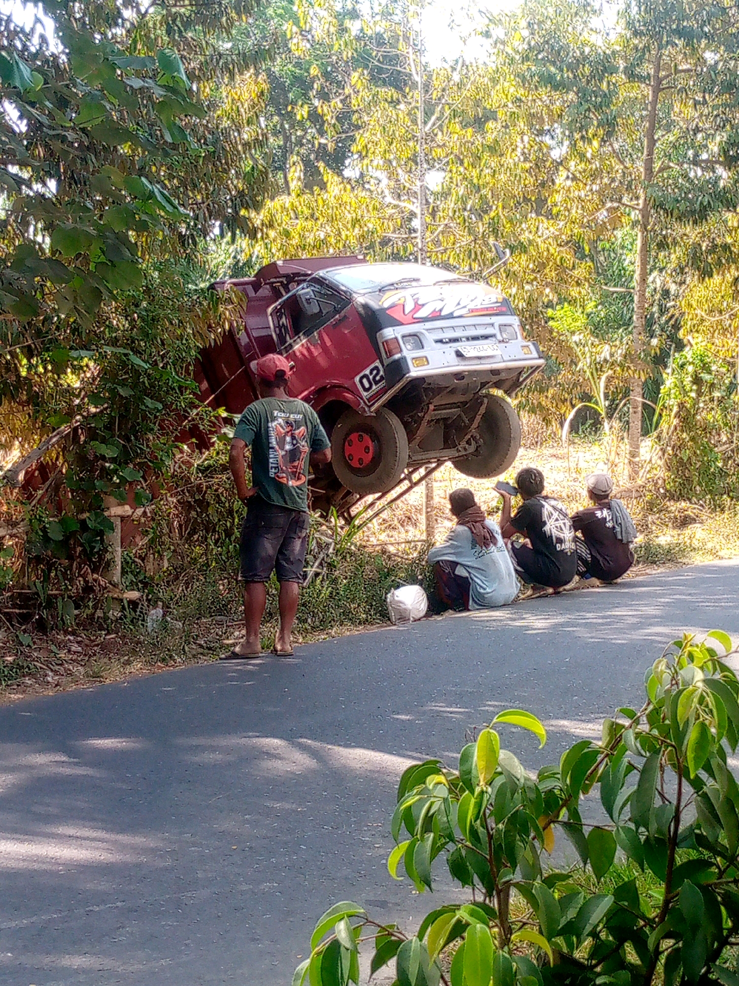 In Sumberjambe, Indonesia, a red truck has crashed, its front end lodged precariously in dense vegetation on the side of an asphalt road. The truck is tilted at a steep angle, with its wheels pointing towards the sky. A man stands on the road, facing away from the truck, observing the scene. Three other individuals are seated on the side of the road, also looking towards the truck. One of them is holding a mobile phone to their ear. The environment is rural, with lush trees and foliage surrounding the road and the crash site. The sun is shining, casting shadows on the road.