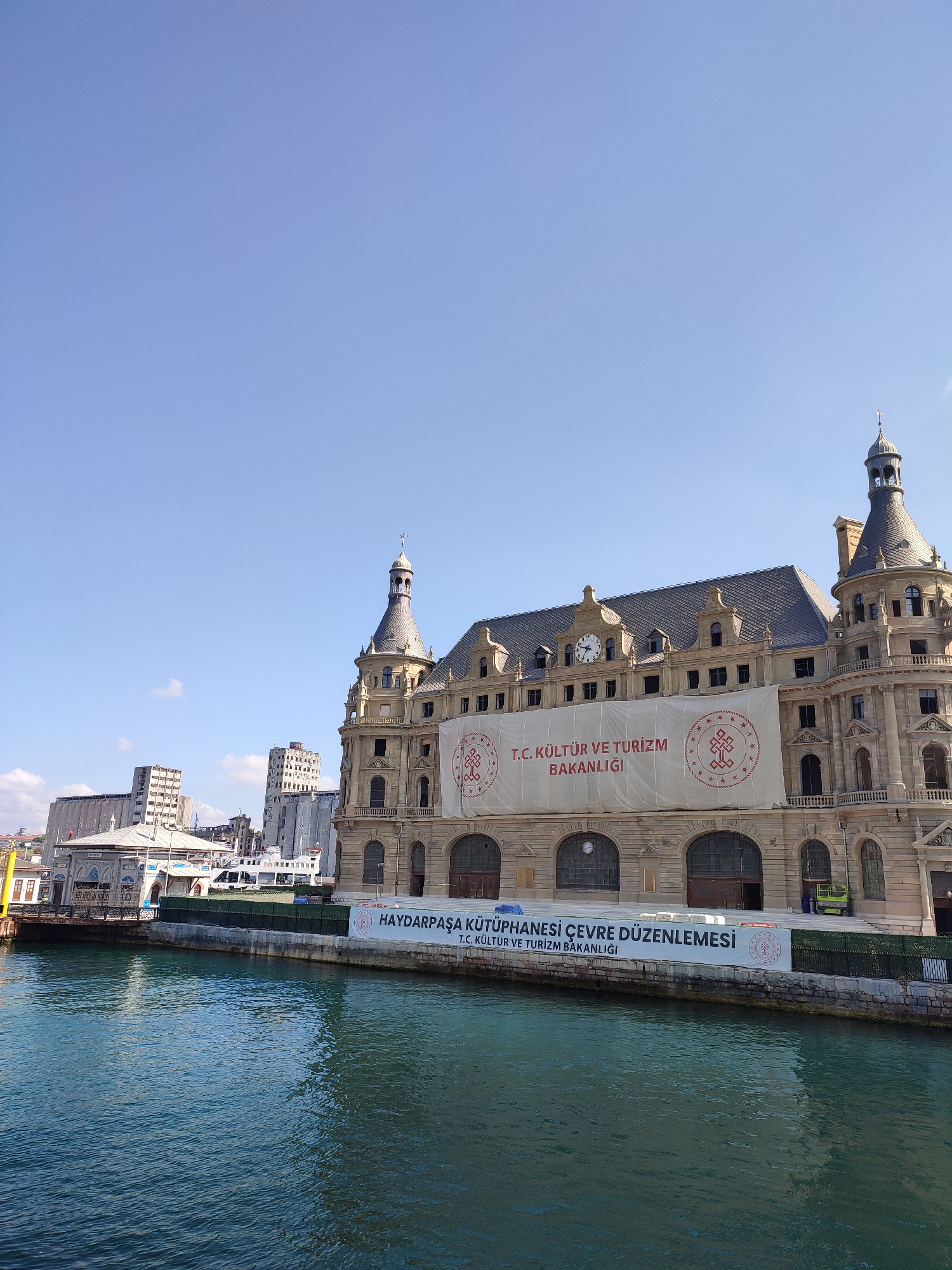An exterior, medium shot captures the Haydarpaşa Train Station in Kadıköy, Turkey, on a clear, bright day. The station, a grand stone structure with multiple towers and a central clock face, is partially obscured by a large white banner. The banner displays text in Turkish, reading "T.C. KÜLTÜR VE TURİZM BAKANLIĞI" and "HAYDARPAŞA KÜTÜPHANESİ ÇEVRE DÜZENLEMESİ". The building is situated on the edge of a large body of water, likely the Bosphorus Strait, which occupies the lower half of the frame. The water is a deep teal color with visible ripples. In the background, a few other modern buildings are visible, including a tall, grey structure and a lighter, more industrial-looking building. The sky is a pale blue with no clouds. There are no people clearly visible in the frame.