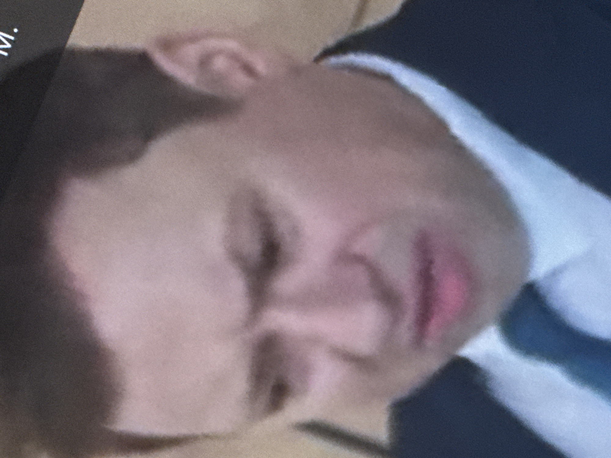 A man is shown from the chest up, his face contorted in a grimace. He is wearing a dark suit jacket and a white collared shirt with a blue tie. The background is blurred and indistinct. The image is grainy and of low resolution.
