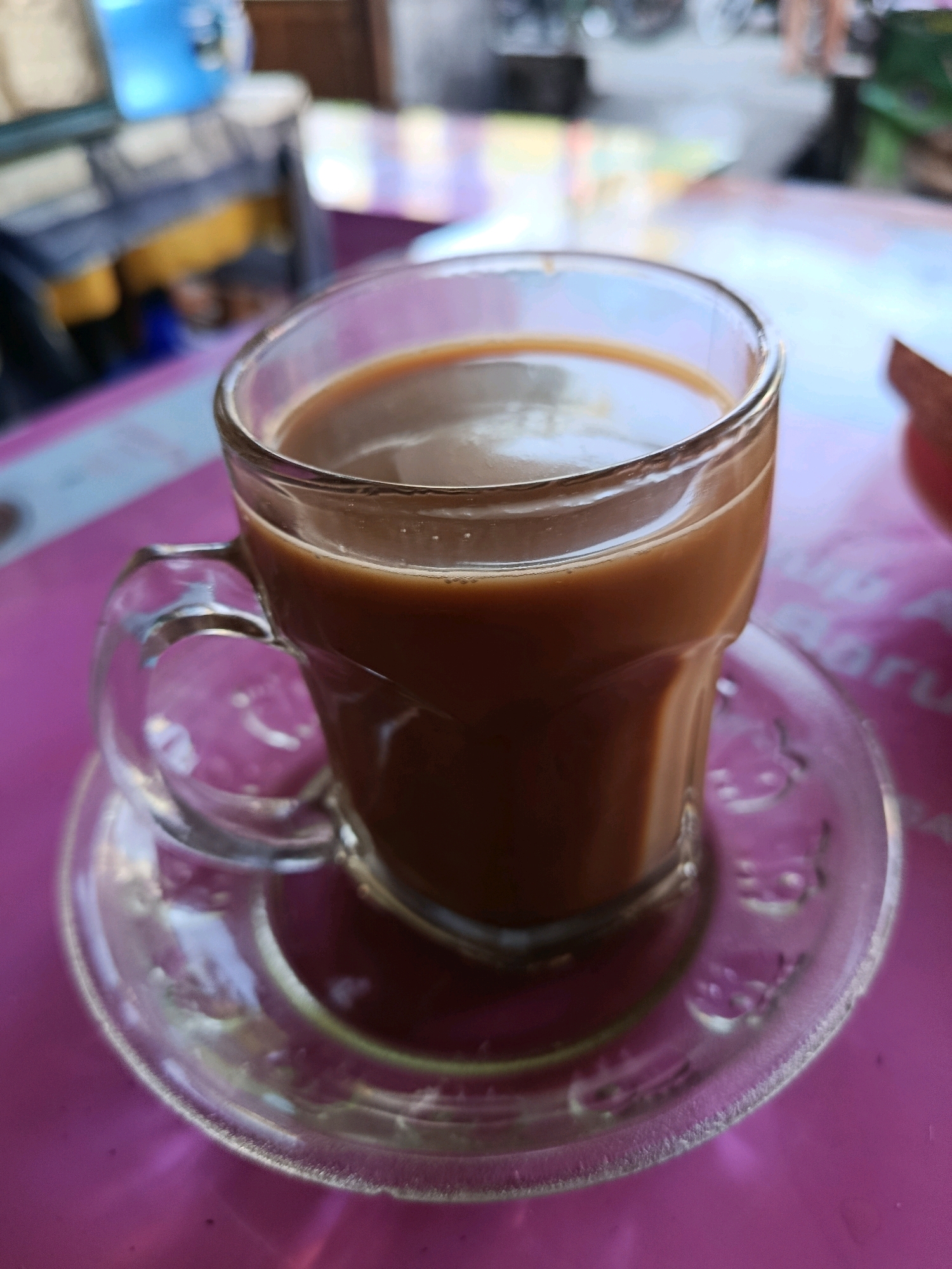 A clear glass mug filled with a brown beverage, likely coffee or tea with milk, sits on a clear glass saucer. The mug has a curved handle. The saucer rests on a textured pink surface, possibly a table. The background is out of focus, showing blurred shapes and colors, including what appears to be furniture and possibly some greenery, suggesting an outdoor or semi-outdoor setting. The scene is lit with natural light, casting soft shadows. No people are visible in the image. The location is specified as Bululawang, Indonesia.