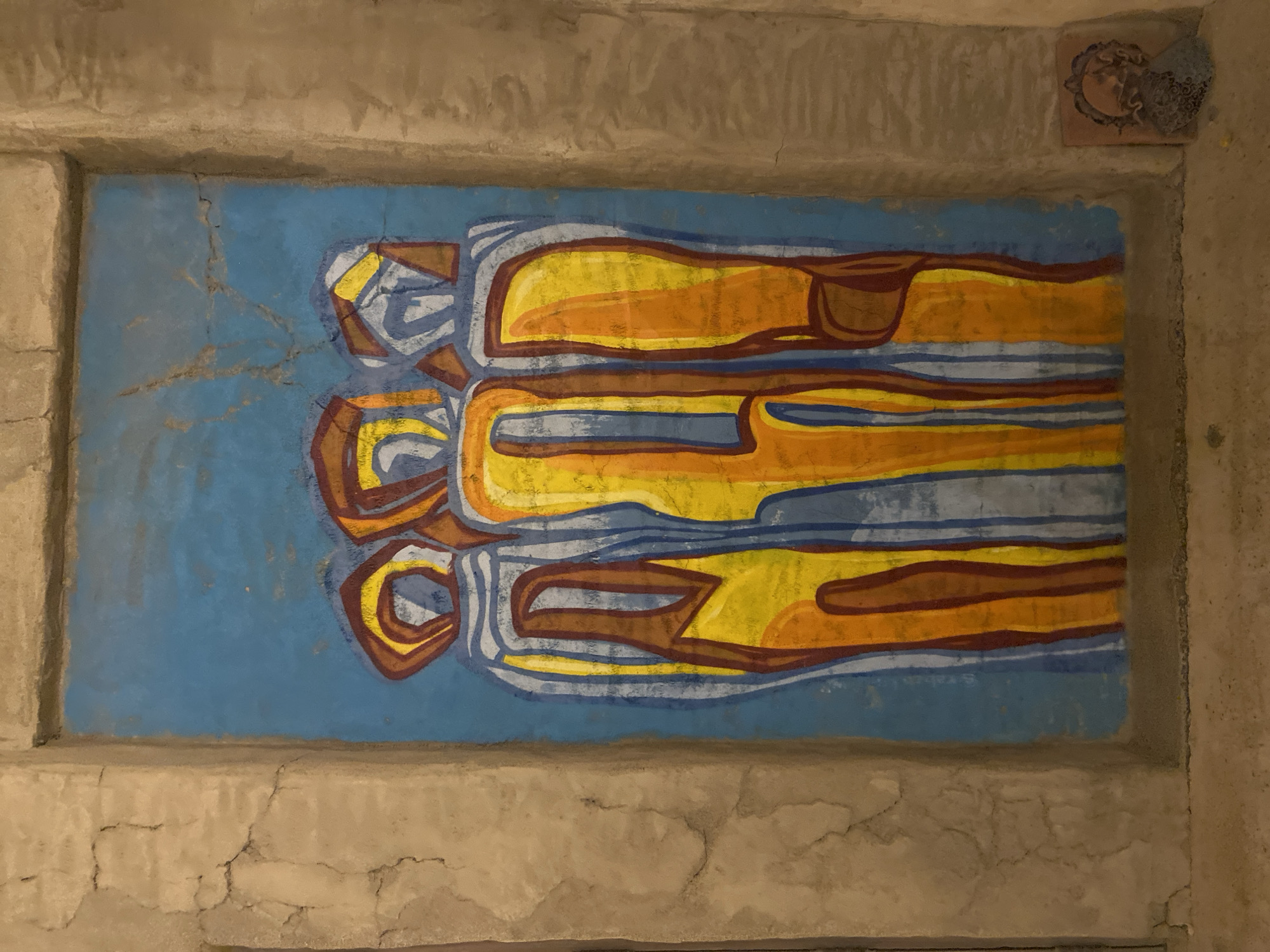 The image shows a mural of three abstract figures on a blue background, set within a rough, textured concrete frame. The figures are depicted in vertical, elongated forms with vibrant colors of yellow, orange, and brown, outlined in blue and white. They appear to be standing side-by-side. To the right of the mural, on the ground, is a small, decorative tile with intricate patterns. The overall environment suggests an outdoor or semi-outdoor space with a rustic, unfinished aesthetic, consistent with the appearance of a wall in a location like Deira, United Arab Emirates, known for its traditional architecture and marketplaces. The lighting is dim, highlighting the textures of the concrete and the colors of the mural.
